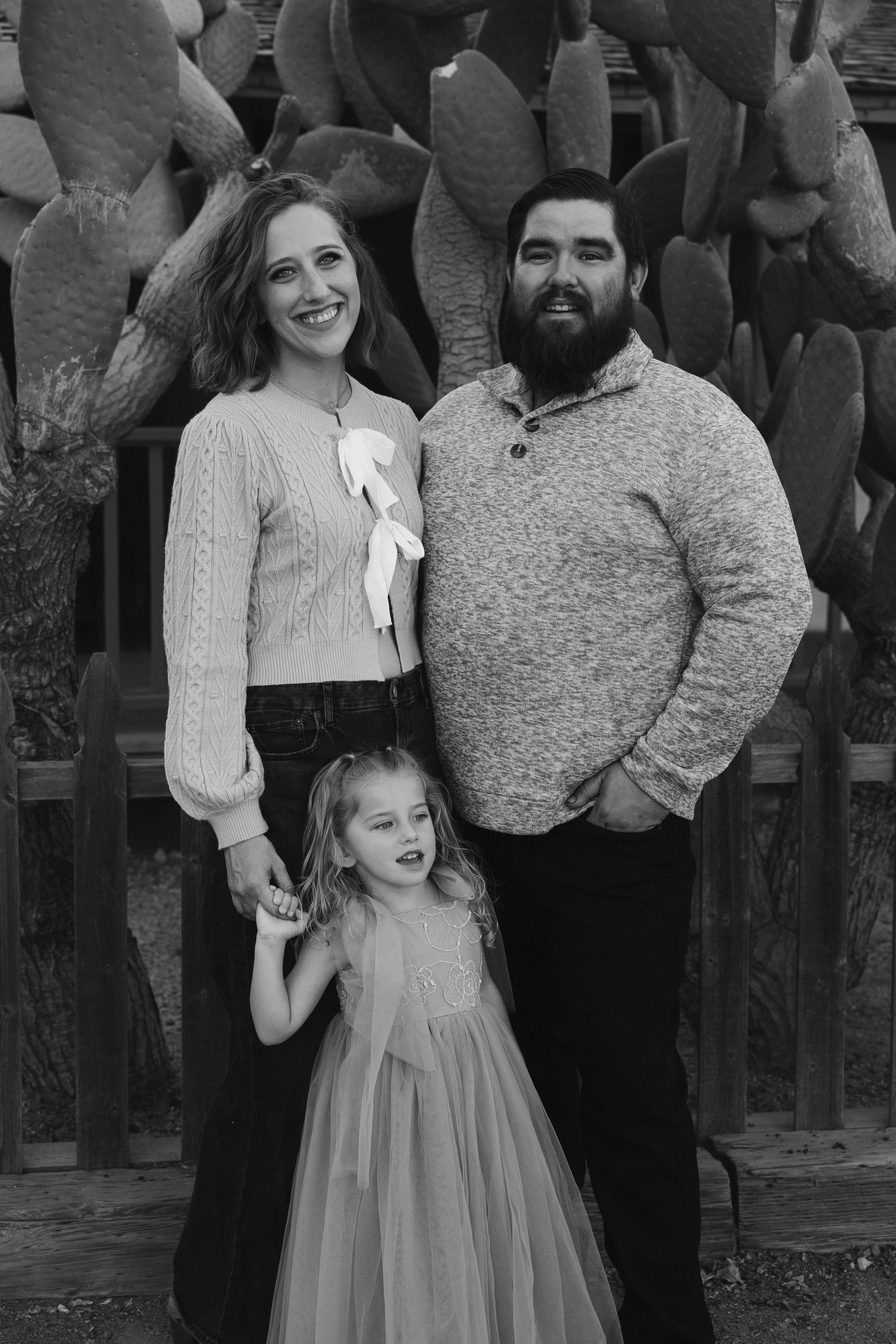 A black-and-white portrait of a family standing in front of large cactus plants, with a woman in a knitted sweater, a bearded man in a textured sweater, and a young girl in a dress holding the woman's hand.