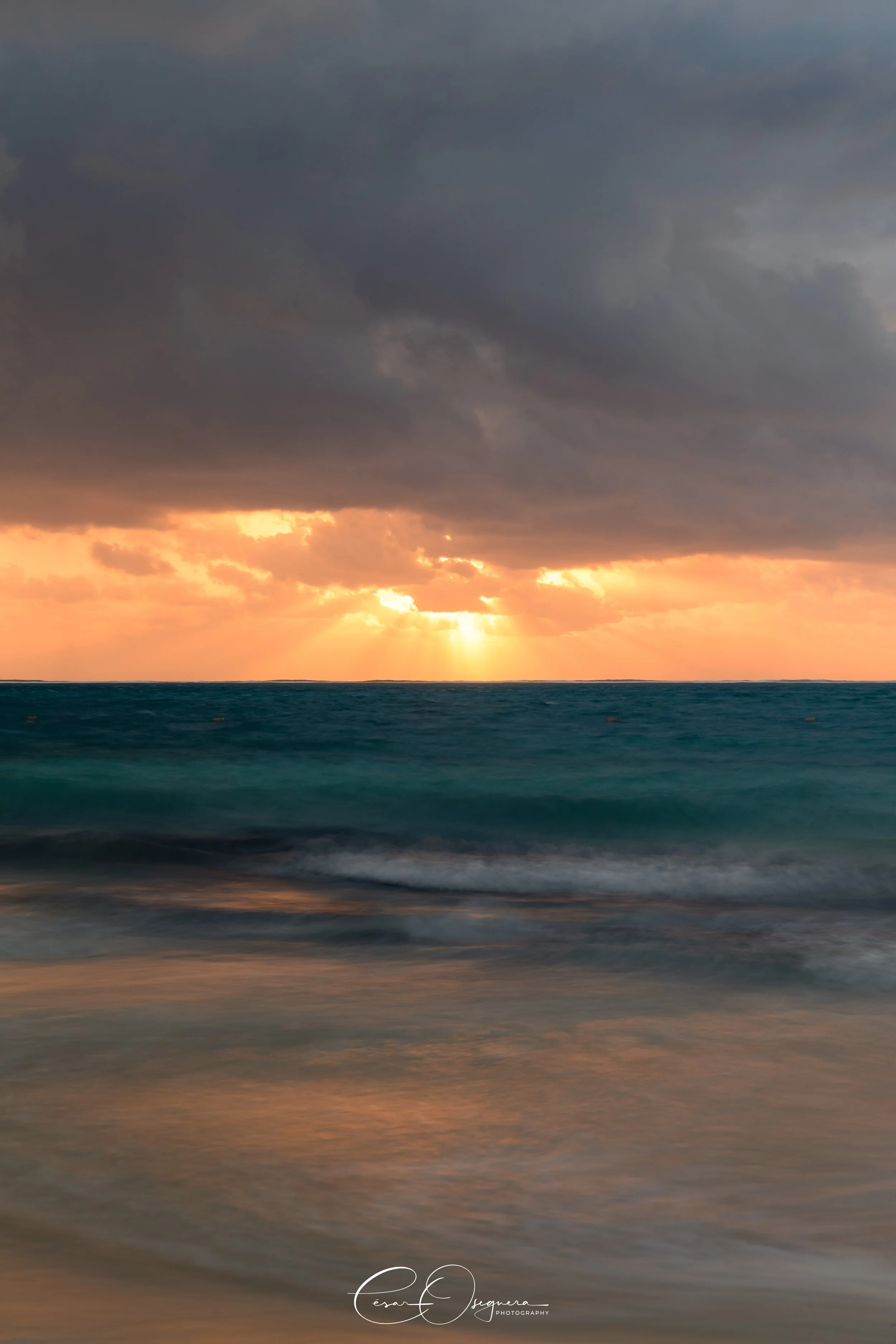 Sunset over the ocean with dark clouds and sun rays breaking through, reflection on the water and gentle waves on the sandy beach.