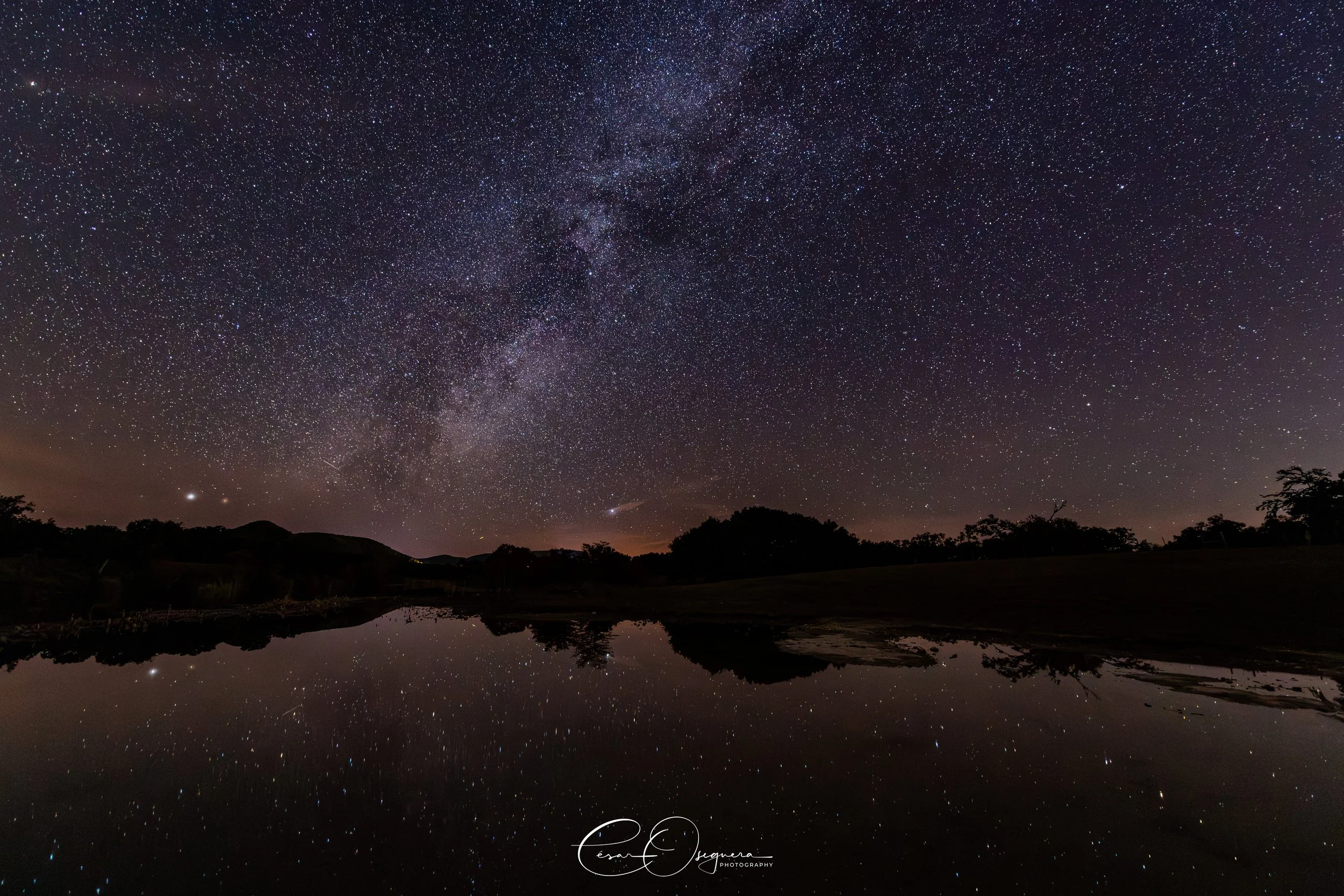 Night sky filled with stars and the Milky Way galaxy above a body of water reflecting the stars, with silhouetted trees and hills.
