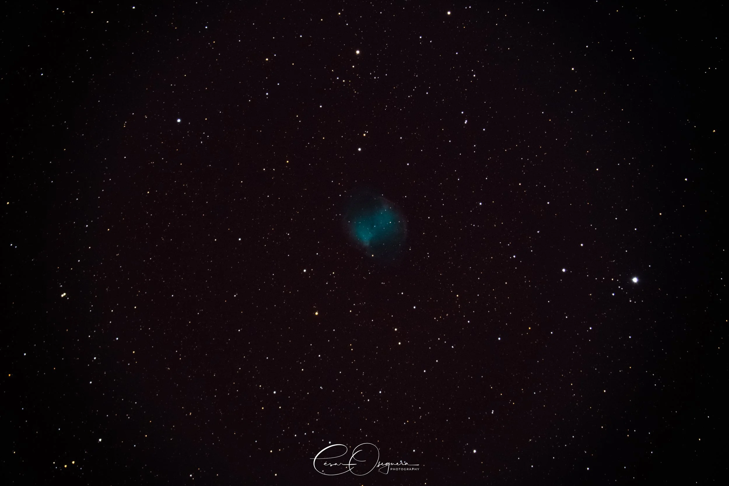A clear night sky filled with stars, featuring a faint, bluish nebula near the center.