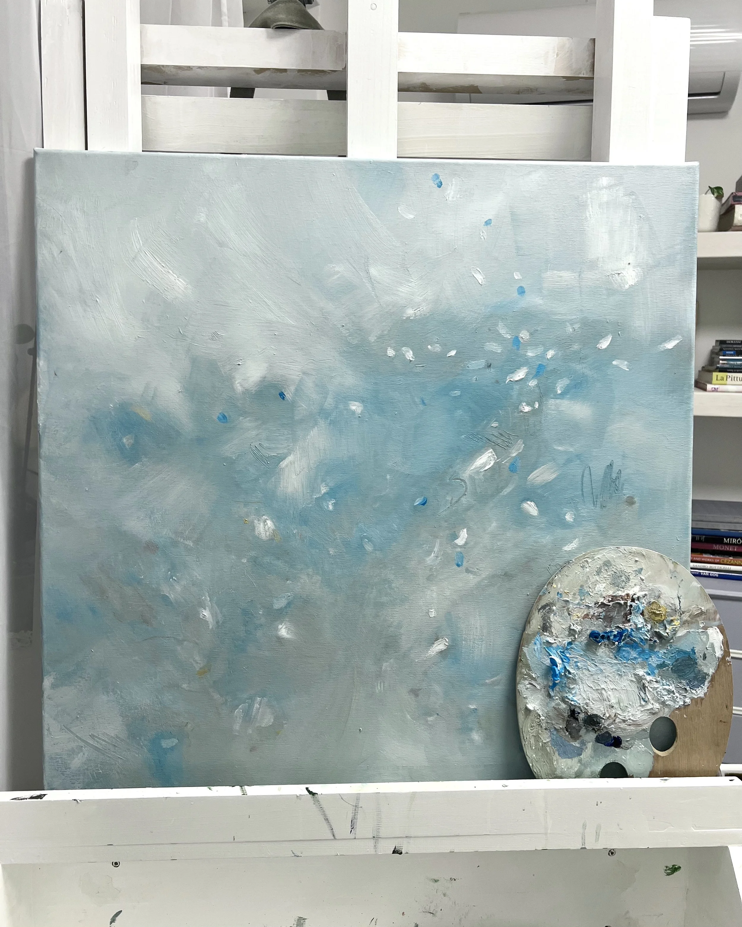 An abstract painting with a predominantly light blue, white, and gray color palette, resting on an easel with a painter's palette filled with mixed paint in front of it. The background includes a white shelf with books and a plant.