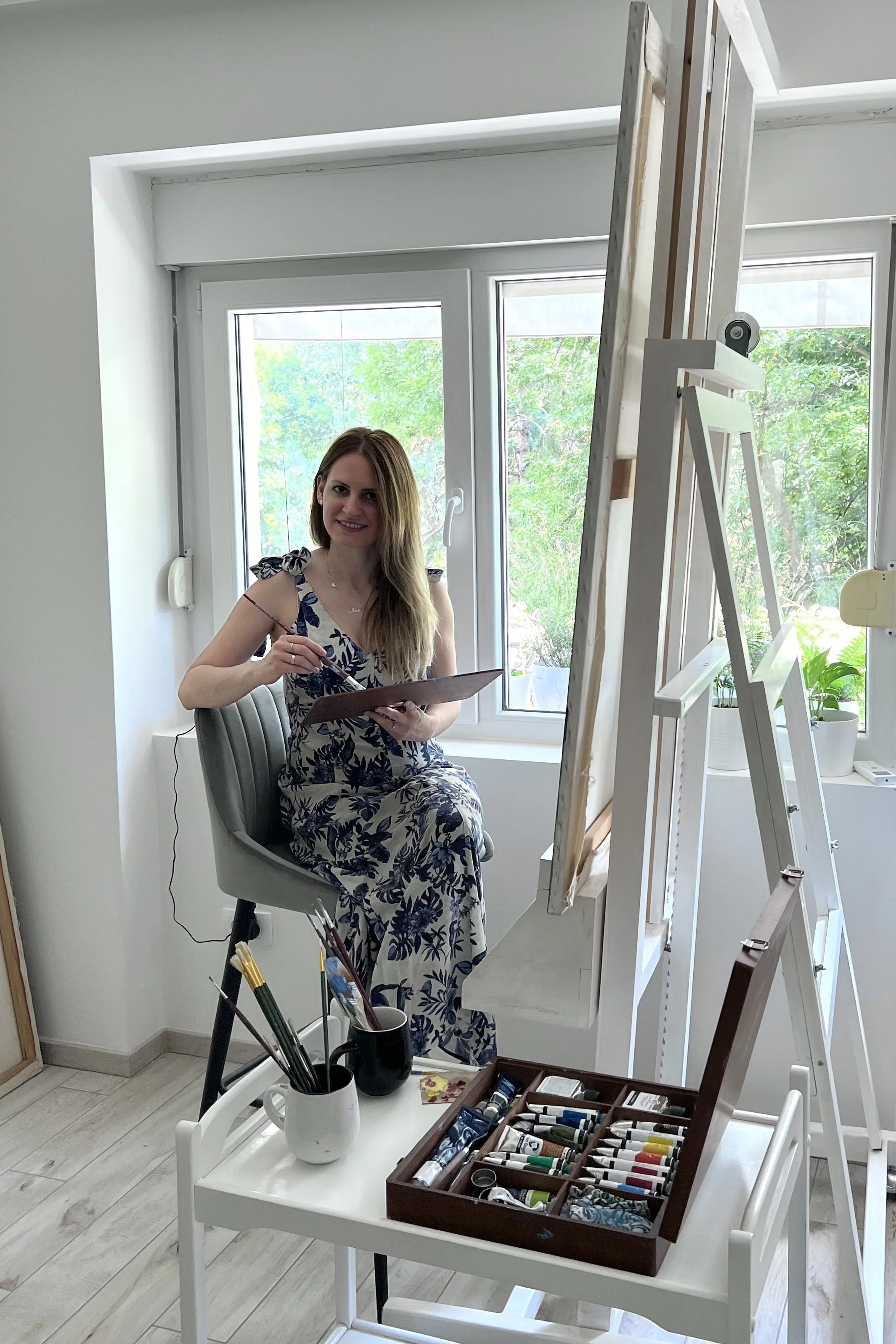 Woman artist sitting on a stool, painting on a large canvas set up on an easel near a window with greenery outside, surrounded by painting supplies, including tubes of paint, brushes, and a palette.