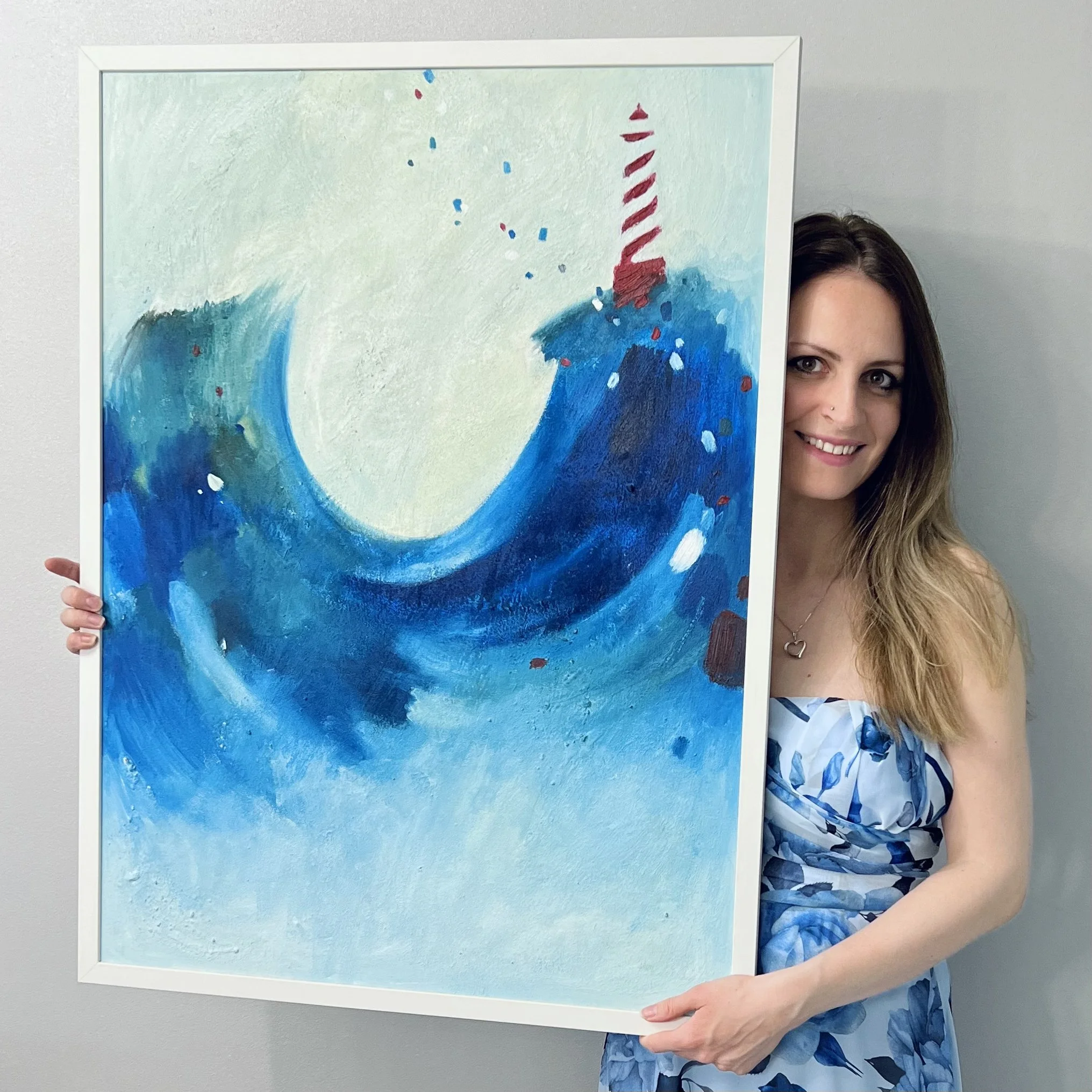 A woman with long brown hair smiling and wearing a blue and white patterned dress, holding a framed abstract painting that features shades of blue, white, red, and teal resembling a wave and possibly a lighthouse in the background.
