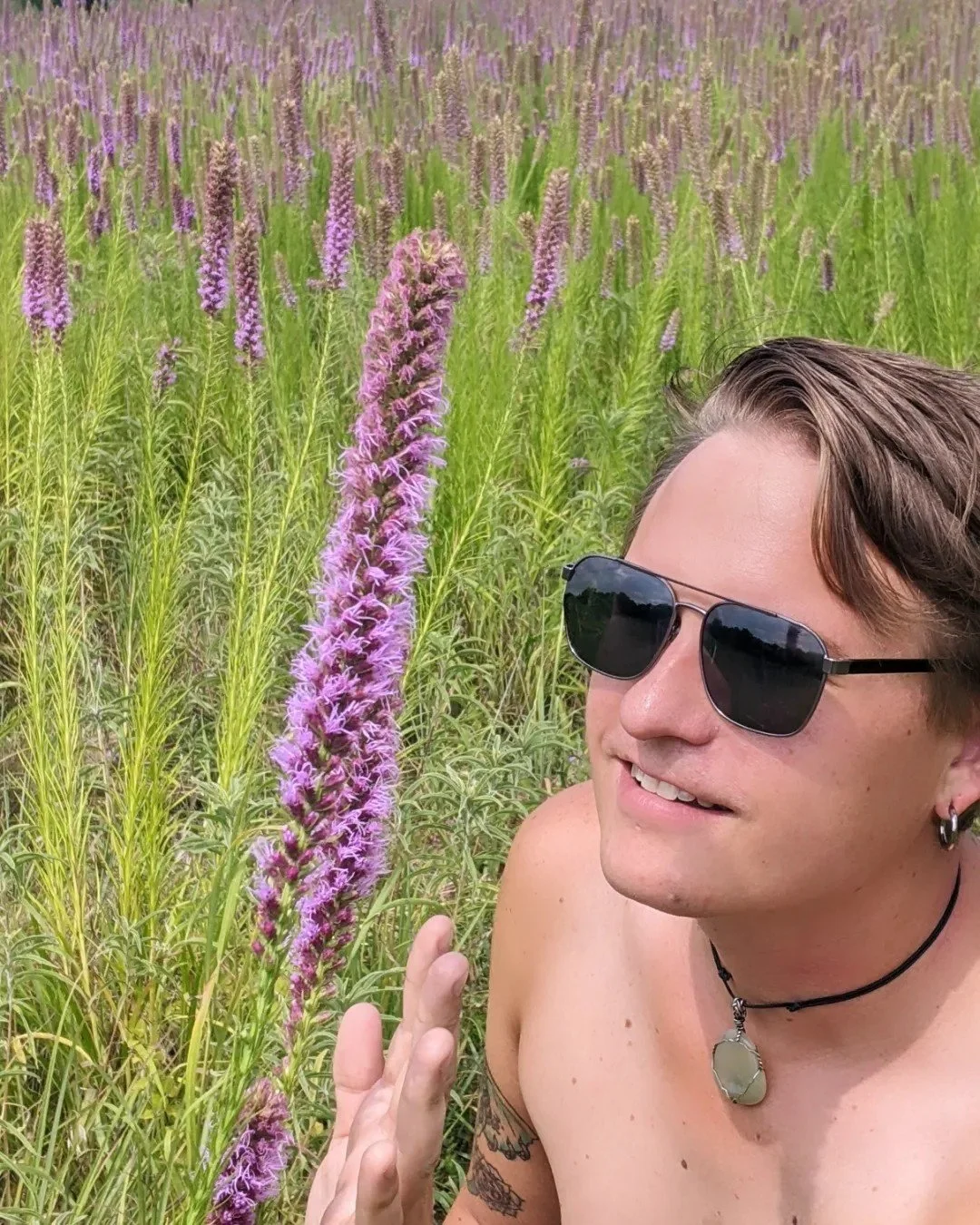 A shirtless  Virgo man with sunglasses, earrings, and a necklace, smiling and reaching out towards tall purple flowers, enjoying nature in a green field of lavender.