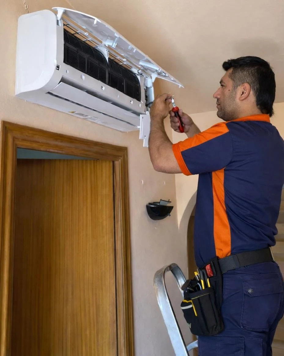 Hombre reparando aire acondicionado con herramientas en una escalera dentro de una casa.