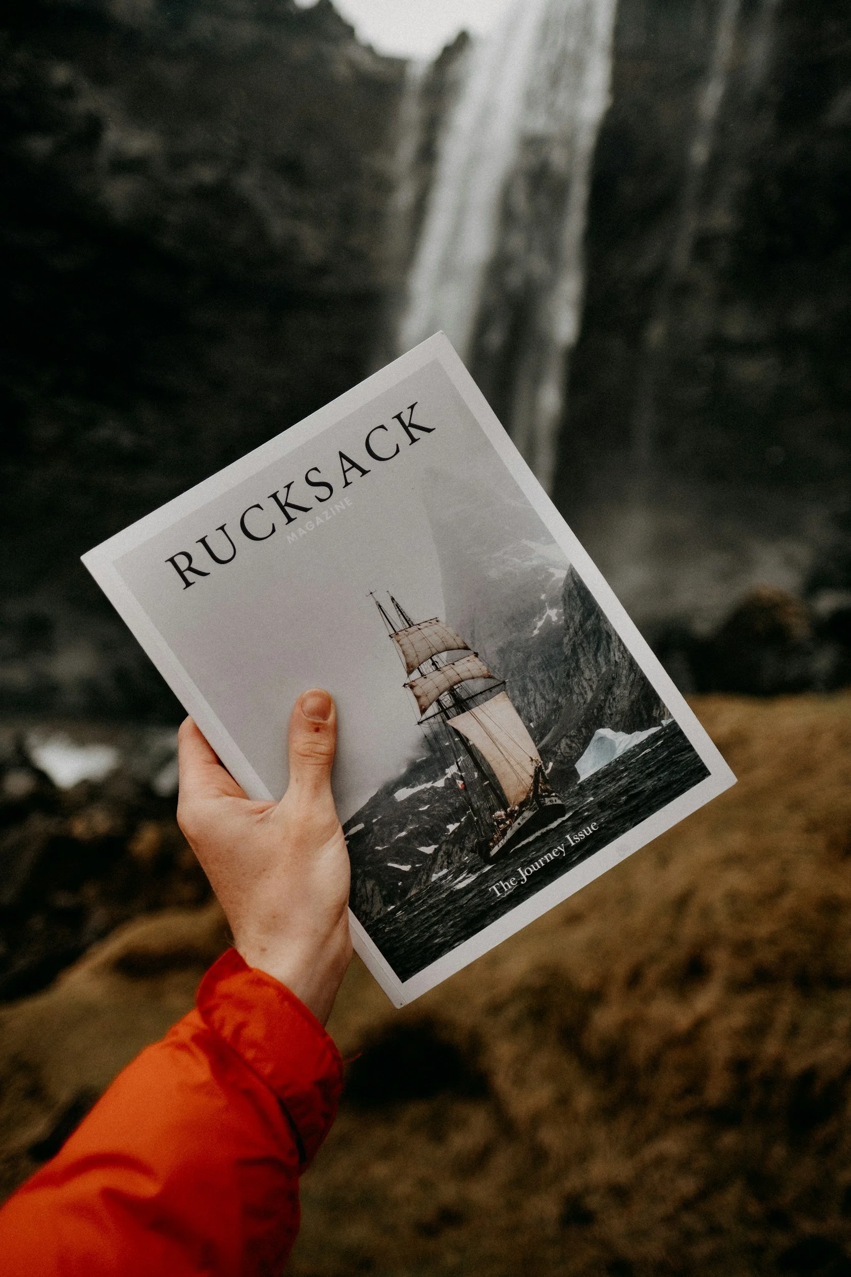 A person holding a magazine titled 'RUCKSACK' with a picture of a sailing ship on the cover, in front of a waterfall.