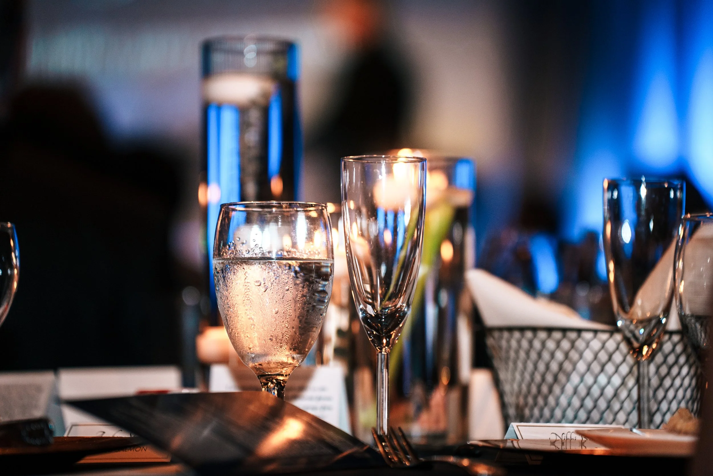 Various glasses on a table with candles in a dimly lit setting, including a water glass and champagne flute, with blurred background.