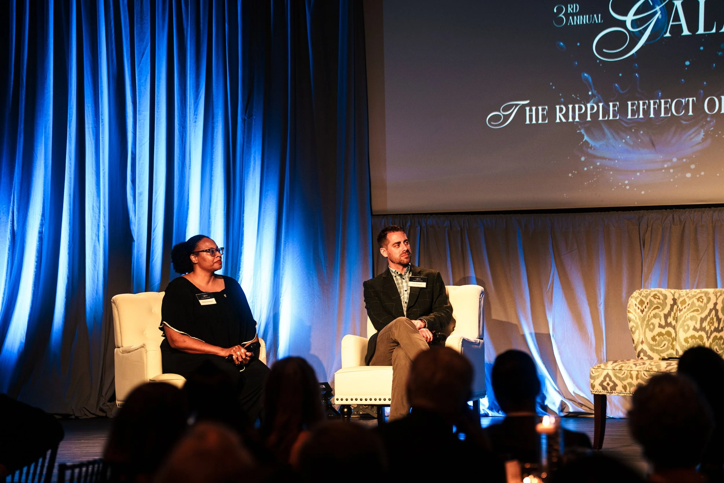 Two people sitting on stage in front of a large screen at a conference or panel discussion, with an audience in front. The screen displays the words '3rd Annual Gala' and 'The Ripple Effect of'. Blue curtains are on the left side of the stage.