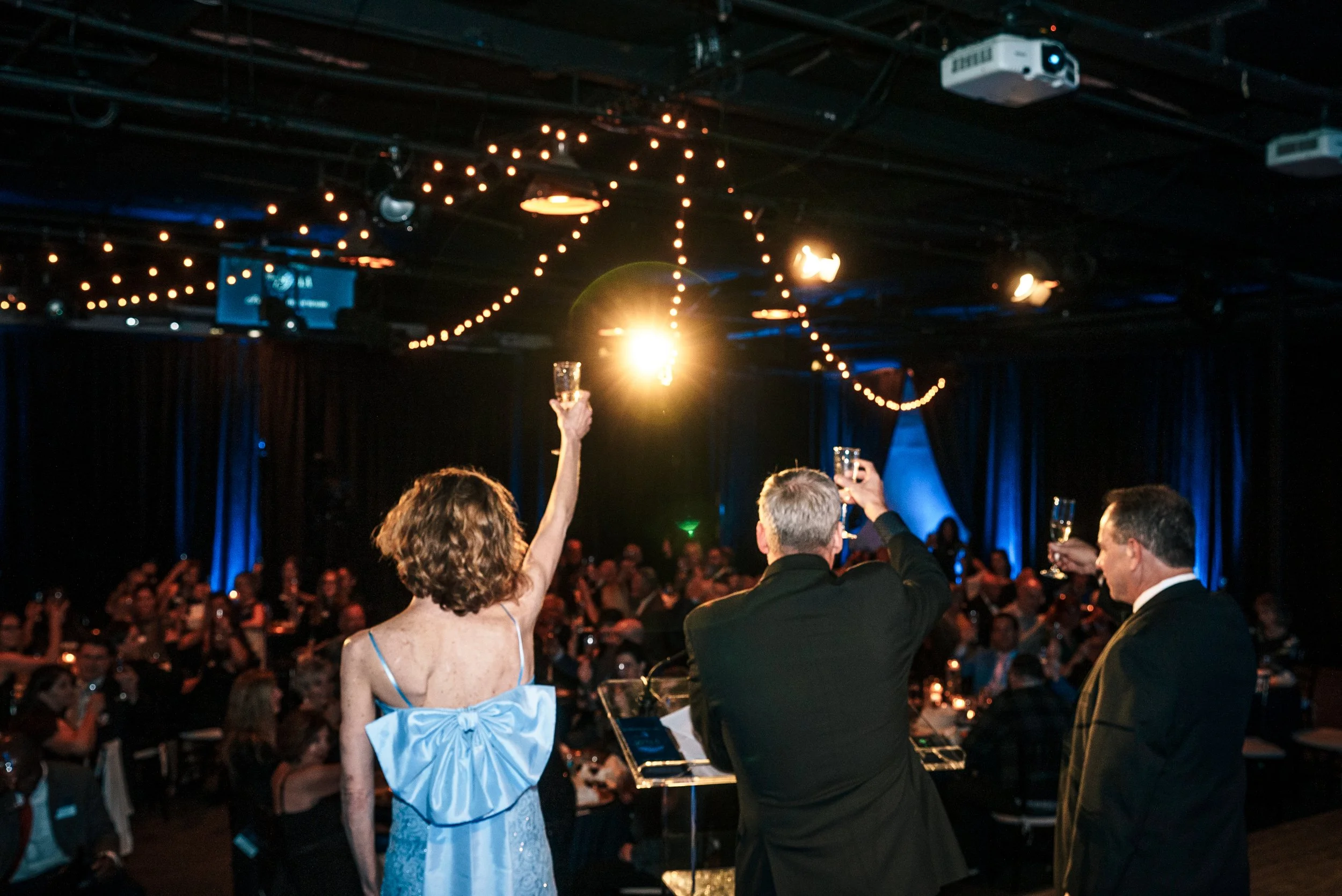 Three people on stage toasting with champagne glasses at a formal event with a seated audience in the background, festive string lights overhead, and a dark atmosphere with blue and orange lighting.
