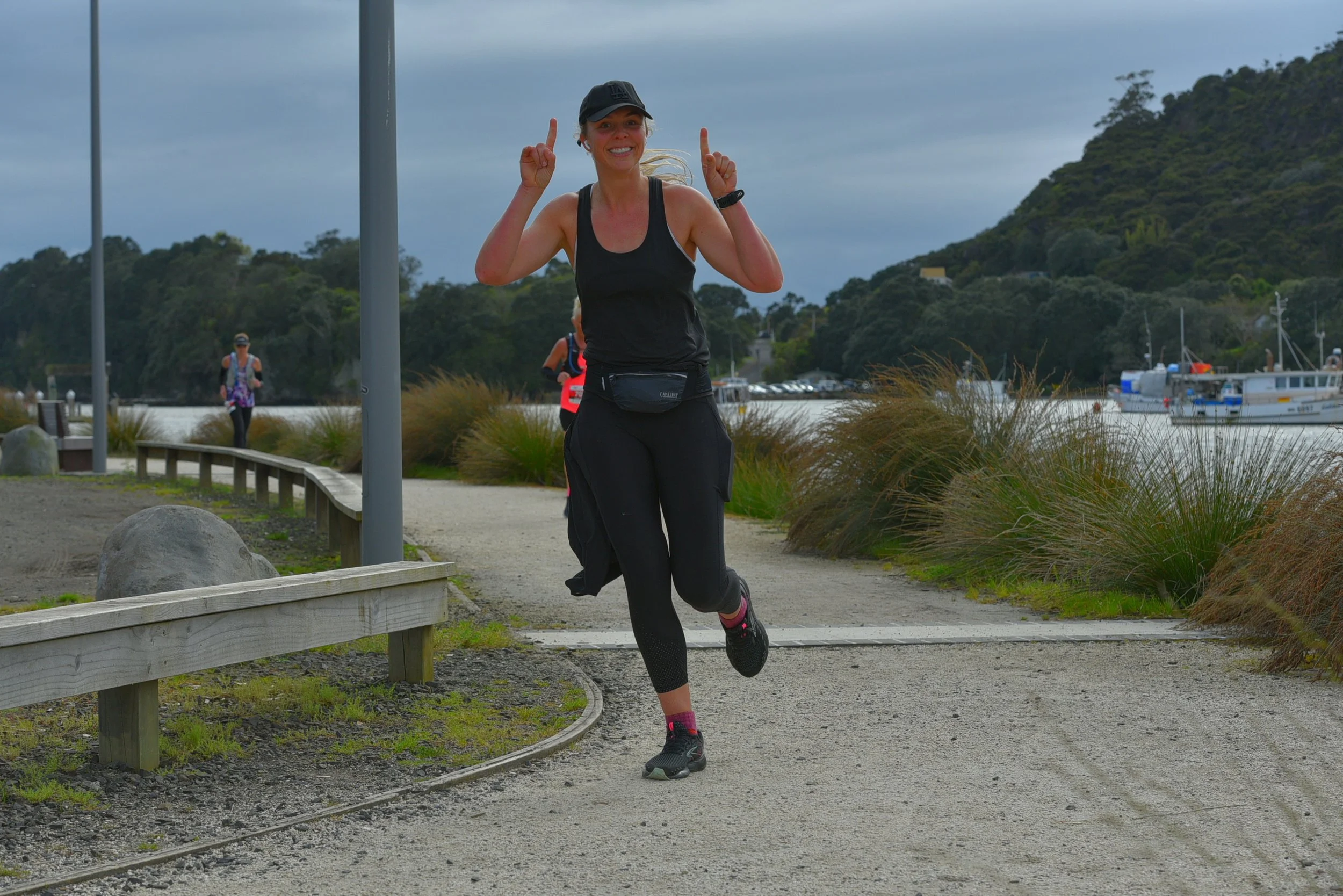Woman jogging on a scenic waterfront path, wearing black athletic clothes and a cap. She has a fanny pack and is pointing upwards with both hands, smiling.