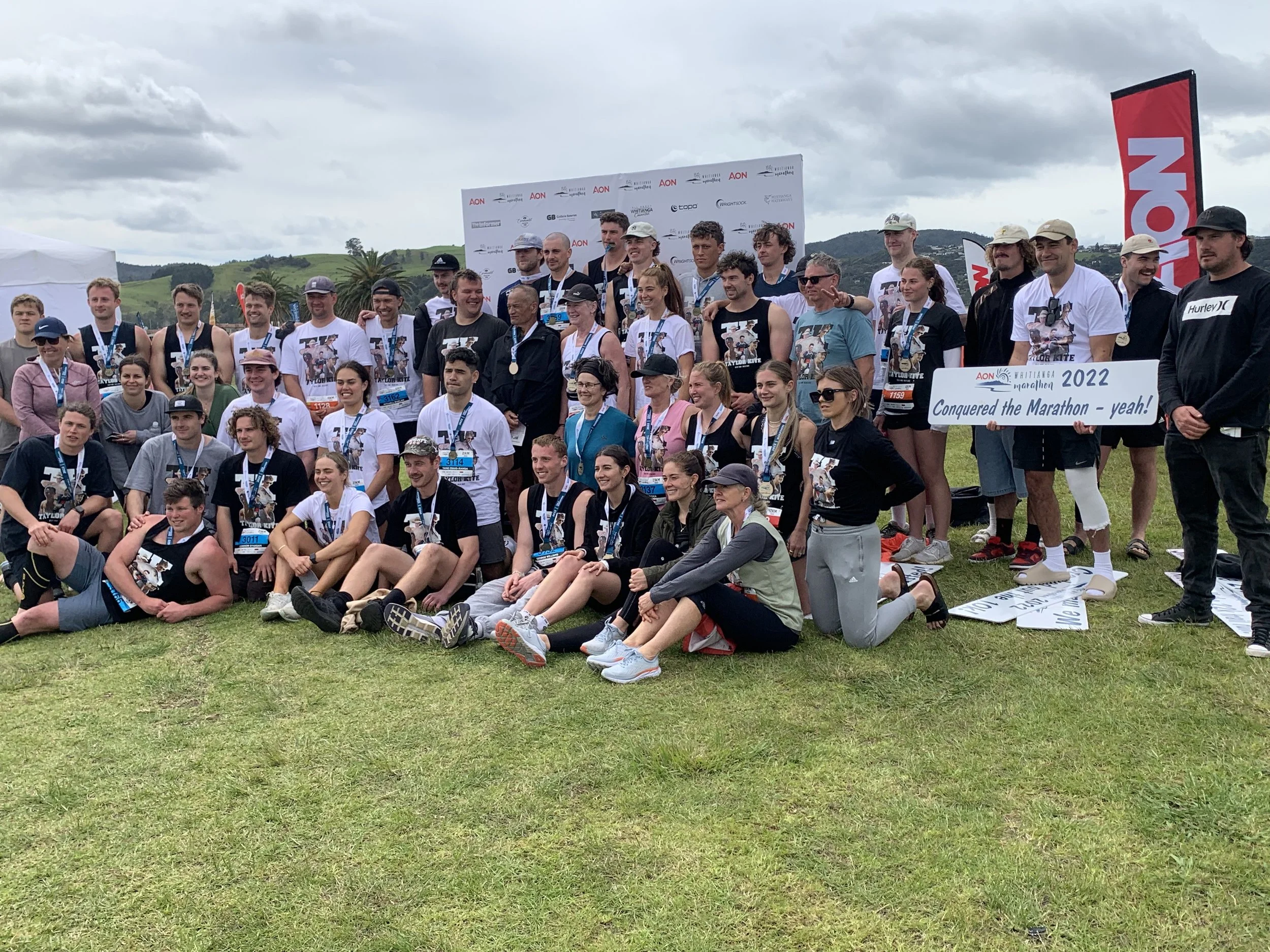 A large group of people posing outdoors on a grassy field, holding a sign that reads "Conquered the Marathon - yeah!" and marked "2022." Many are wearing medals, suggesting they participated in a marathon. A backdrop with logos is behind them, and a 