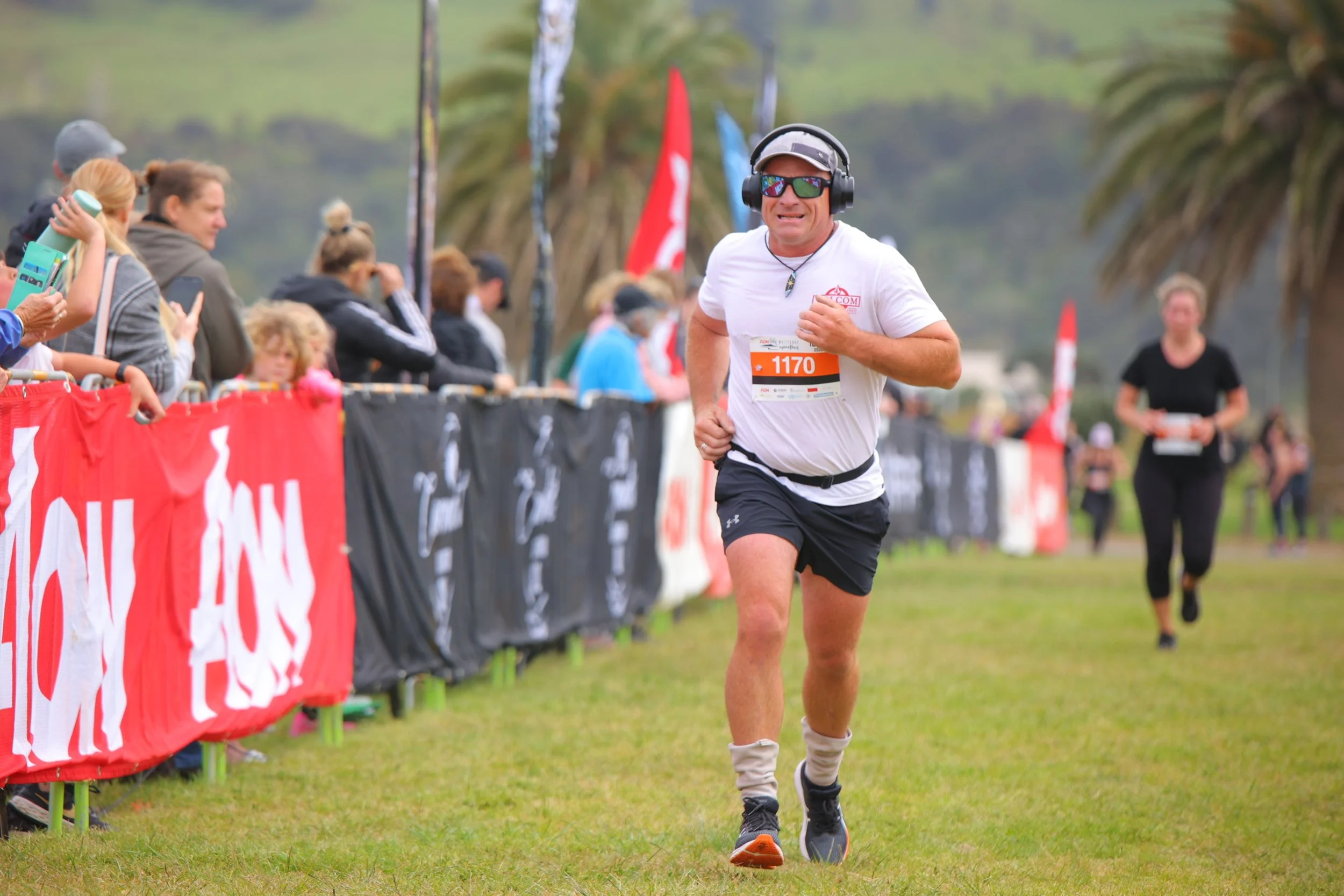 Runner in white shirt and headphones participating in outdoor race, with spectators behind barriers, green grass, and palm trees in background.