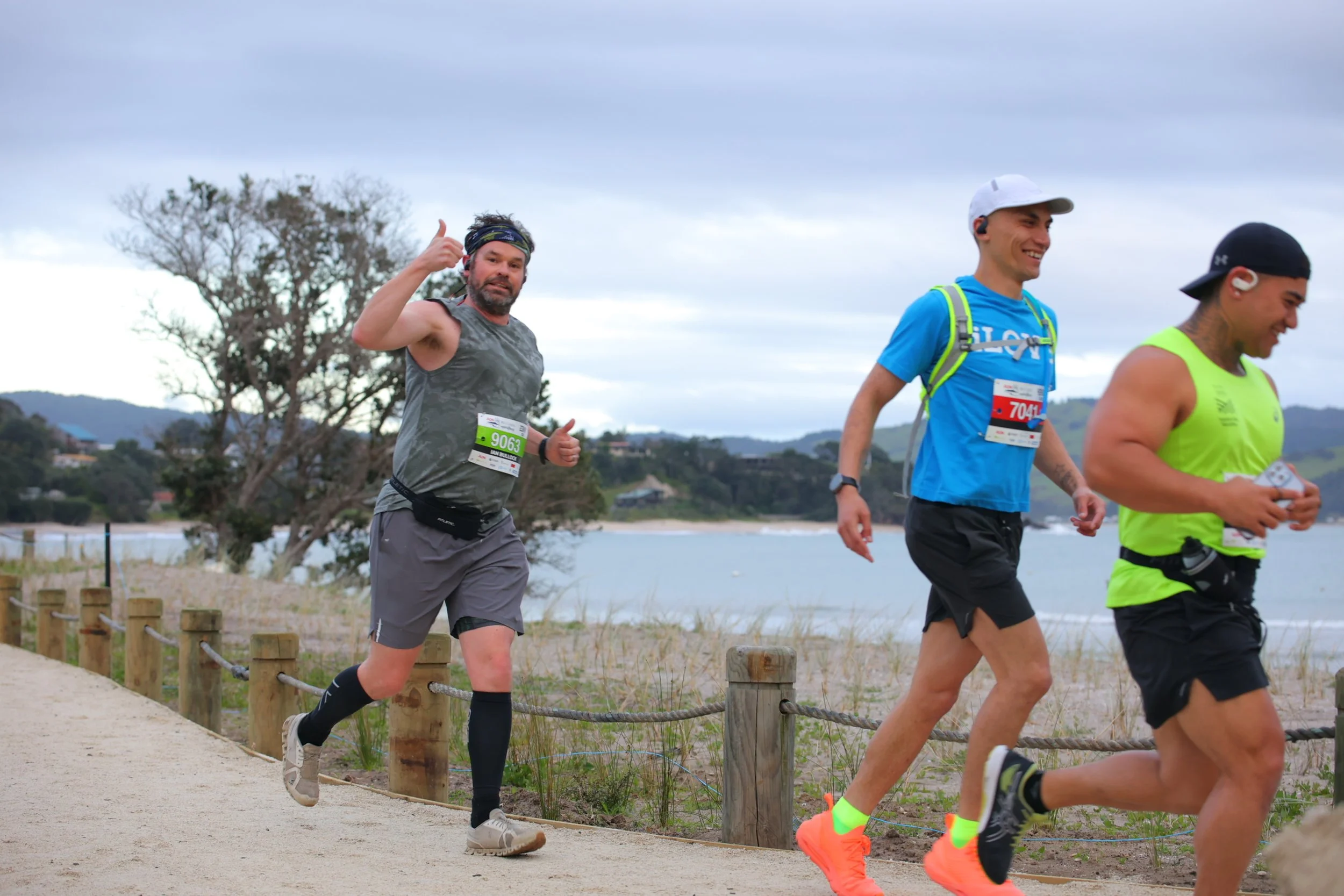 Three men running on a path near a beach, wearing athletic gear and race bibs, with one man giving a thumbs up. Background includes ocean and hills.
