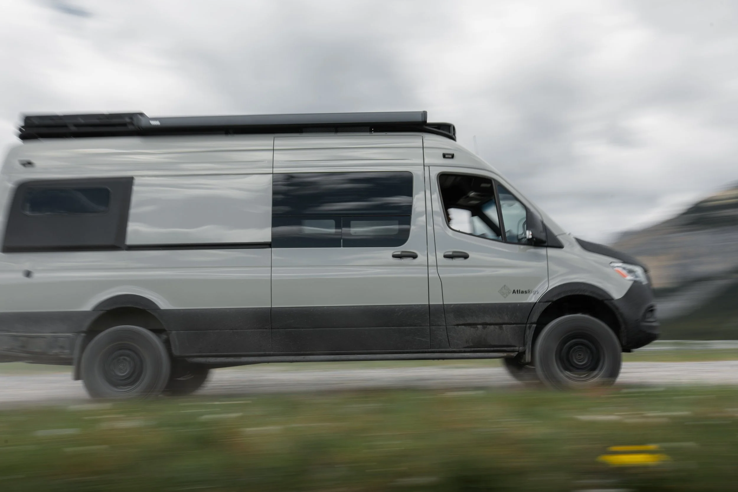 A white camper van with black window accents and a rooftop storage rack, moving on a highway under a cloudy sky.