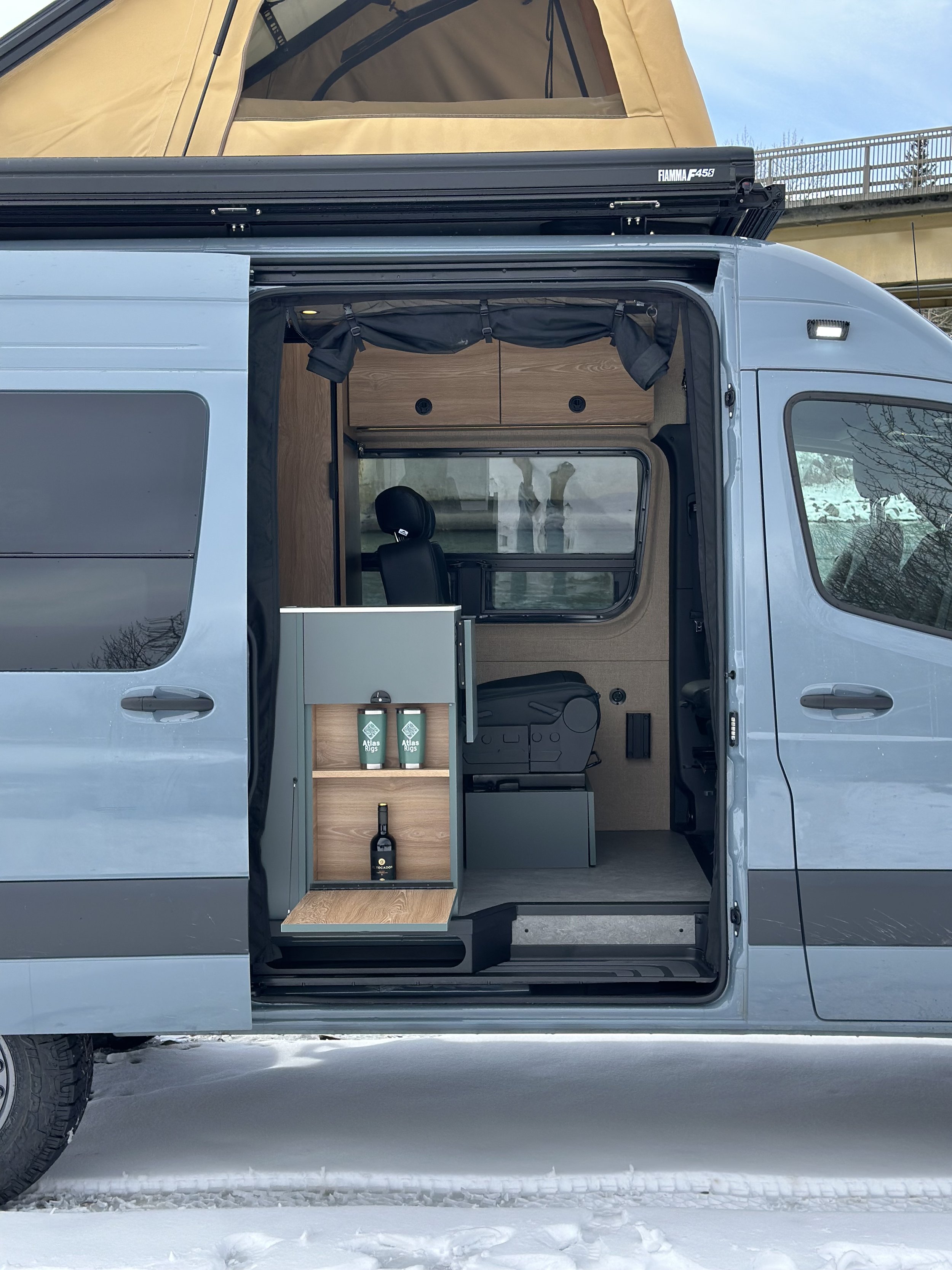 The interior of a camper van with a wooden cabinet, a window with black curtains, and a countertop with two green insulated bottles labeled 'Atlas Rigs'. The van is parked on snow, and a tent is mounted on top of the van.