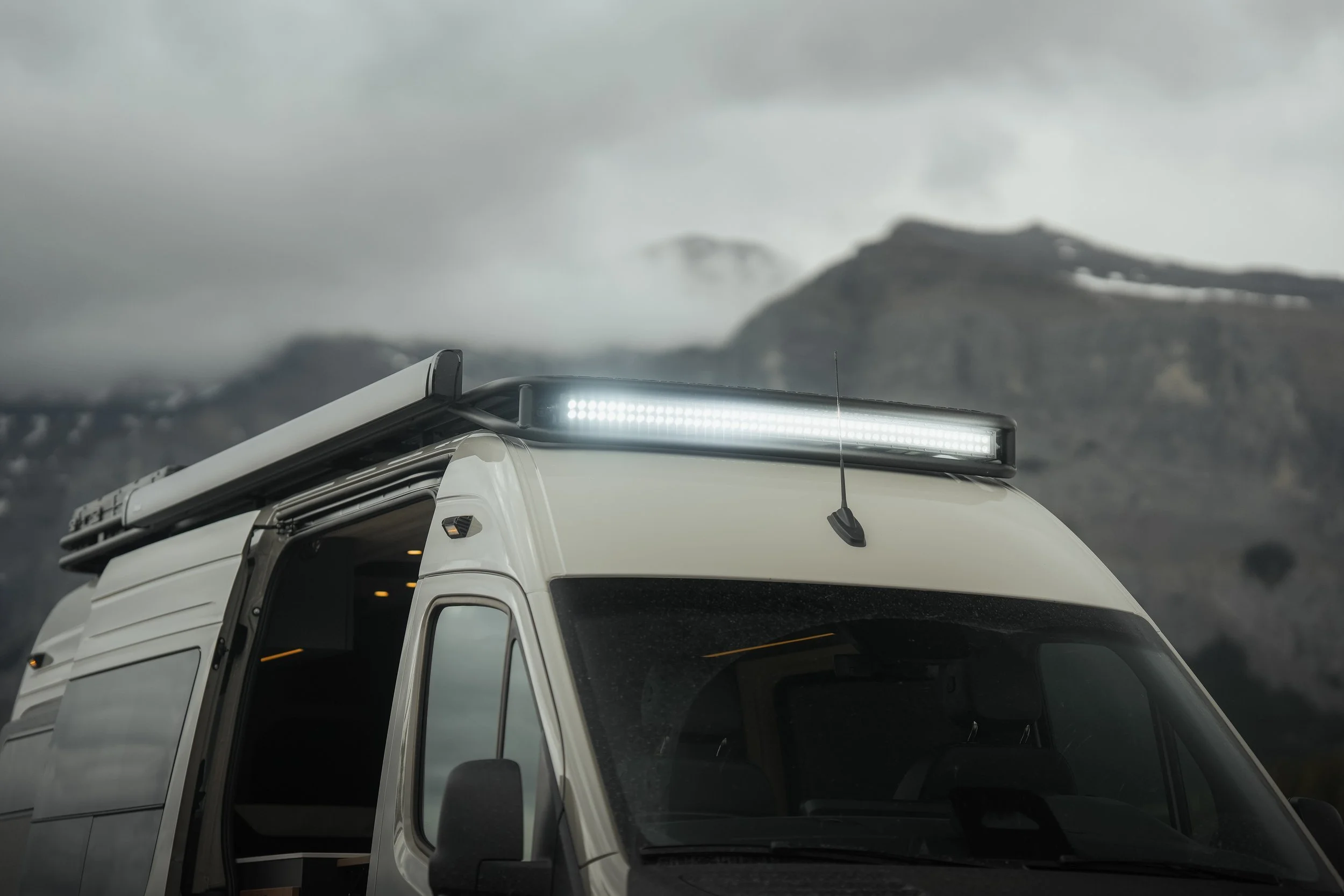 The front part of a white truck with a bright LED light bar on top, against a mountainous, cloudy, and foggy landscape.