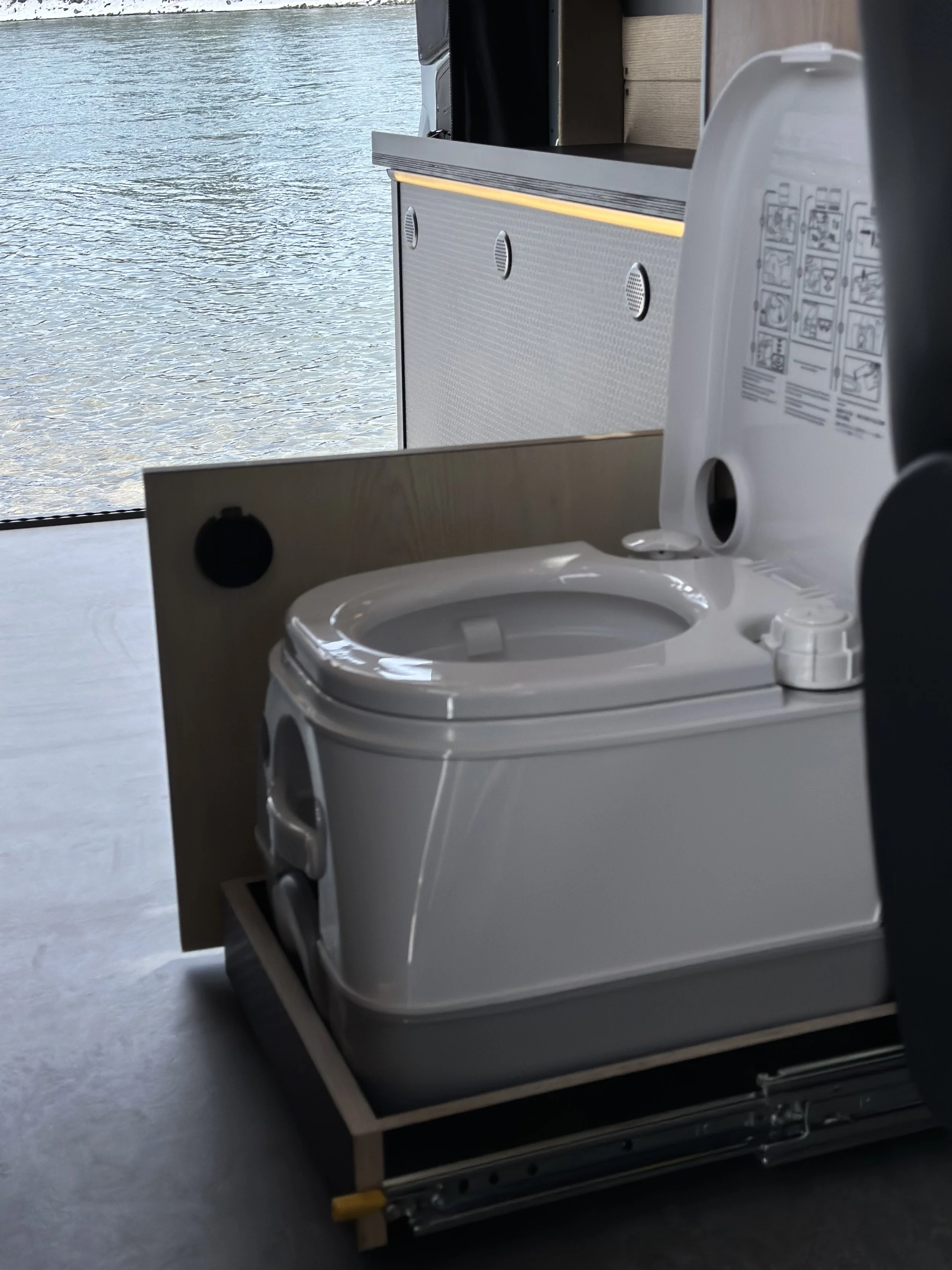 A toilet and a foldable toilet seat in a room with a large window showing water outside.