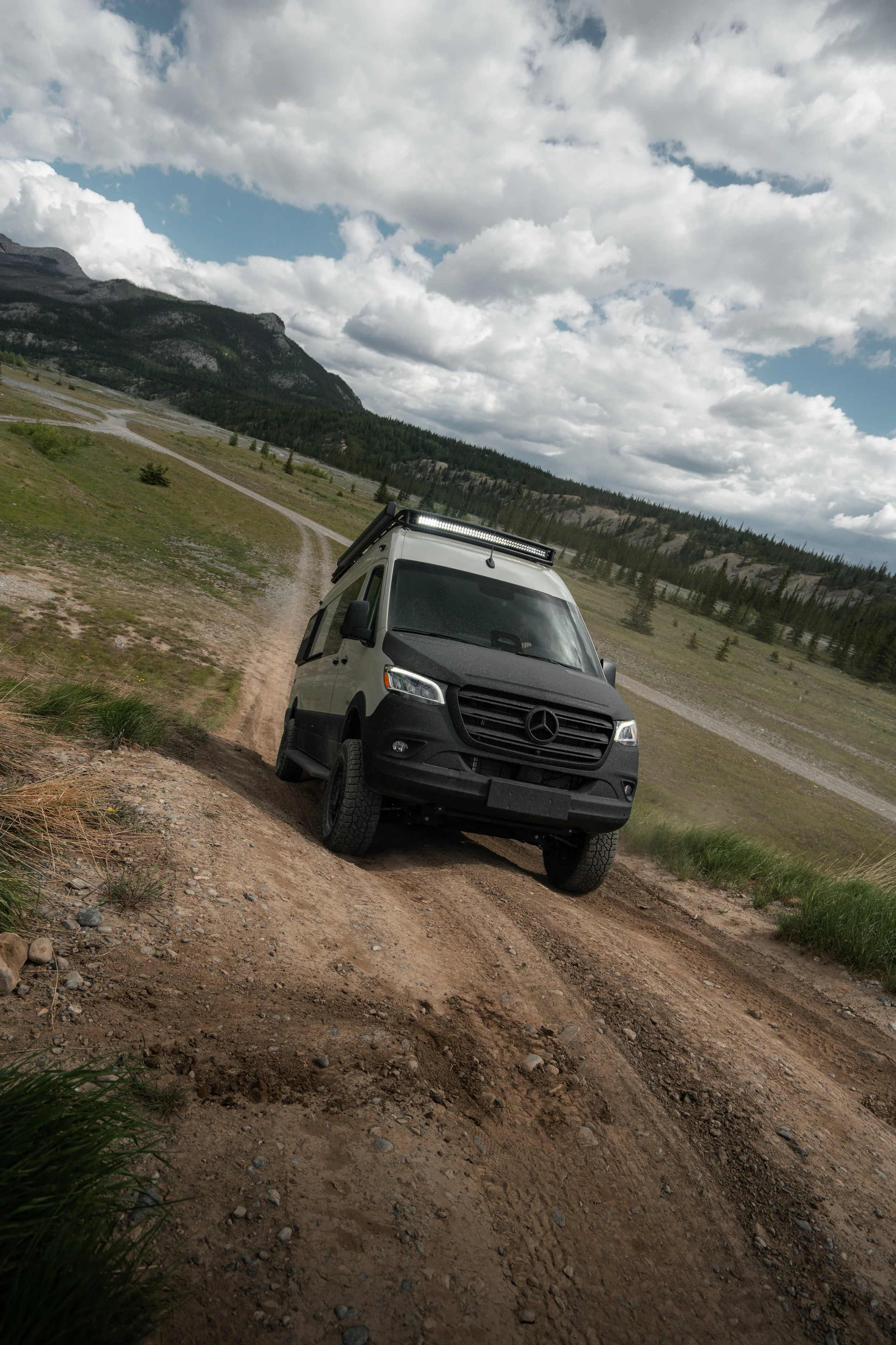 A silver Mercedes-Benz van with off-road tires driving on a dirt trail in a mountainous area under a cloudy sky.