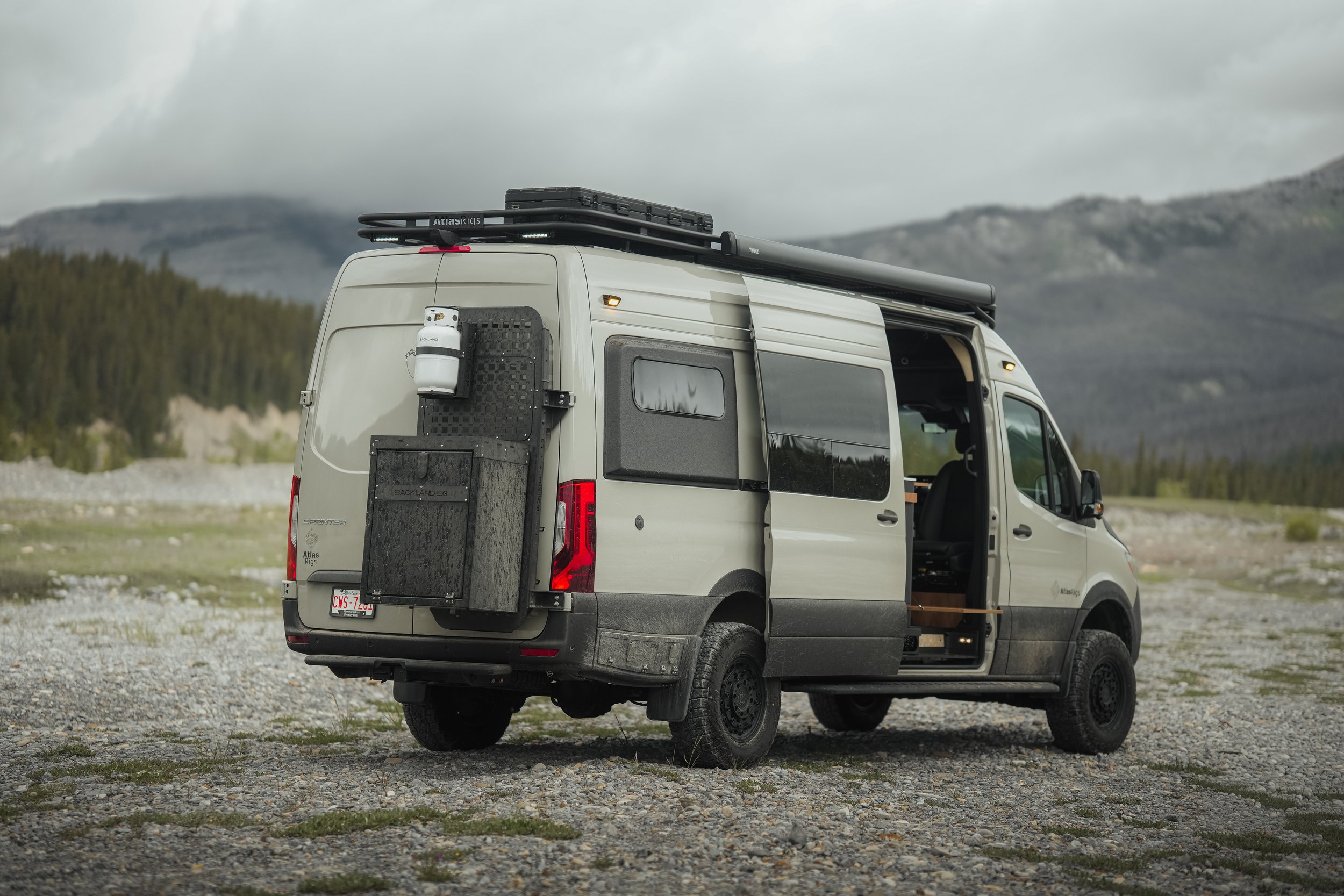 White camper van parked in a mountainous, rocky area with overcast skies, roof rack, and equipment attached to the back.