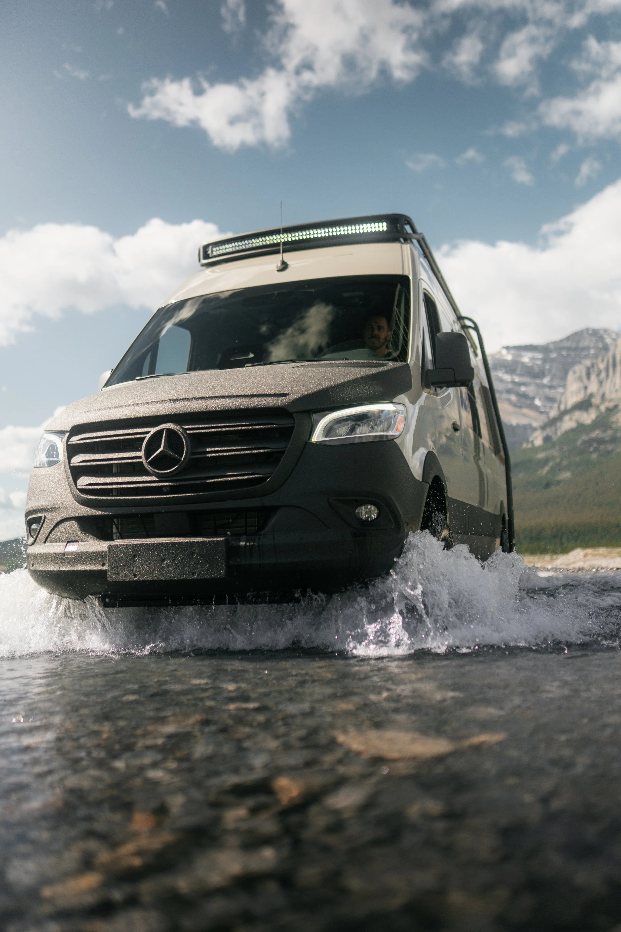 A grey Mercedes-Benz van driving through water with mountains and a blue sky with clouds in the background.