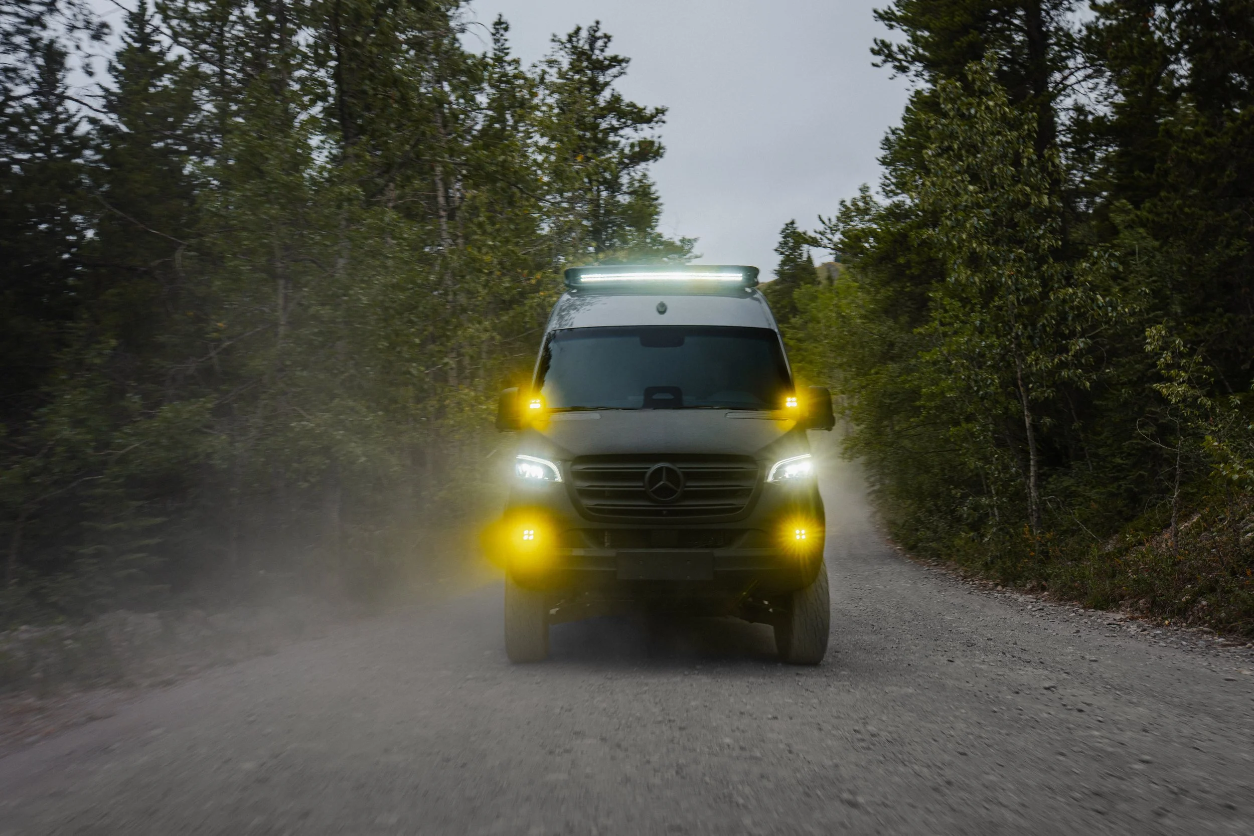 Electric autonomous vehicle with bright LED lights driving on a dirt road through a forested area during daytime.