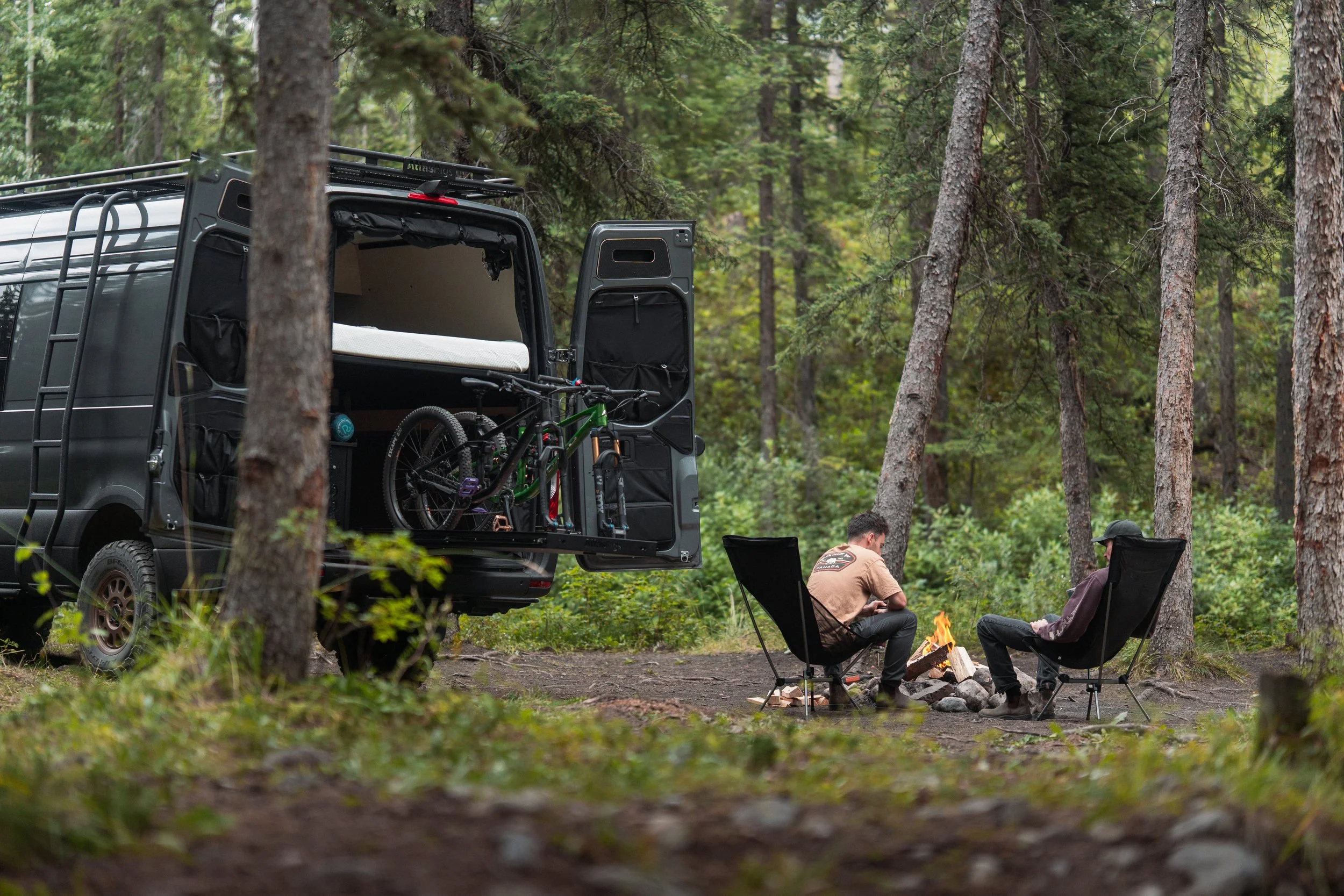 People are relaxing around a campfire in a forest, with an open van nearby carrying bicycles and camping gear.