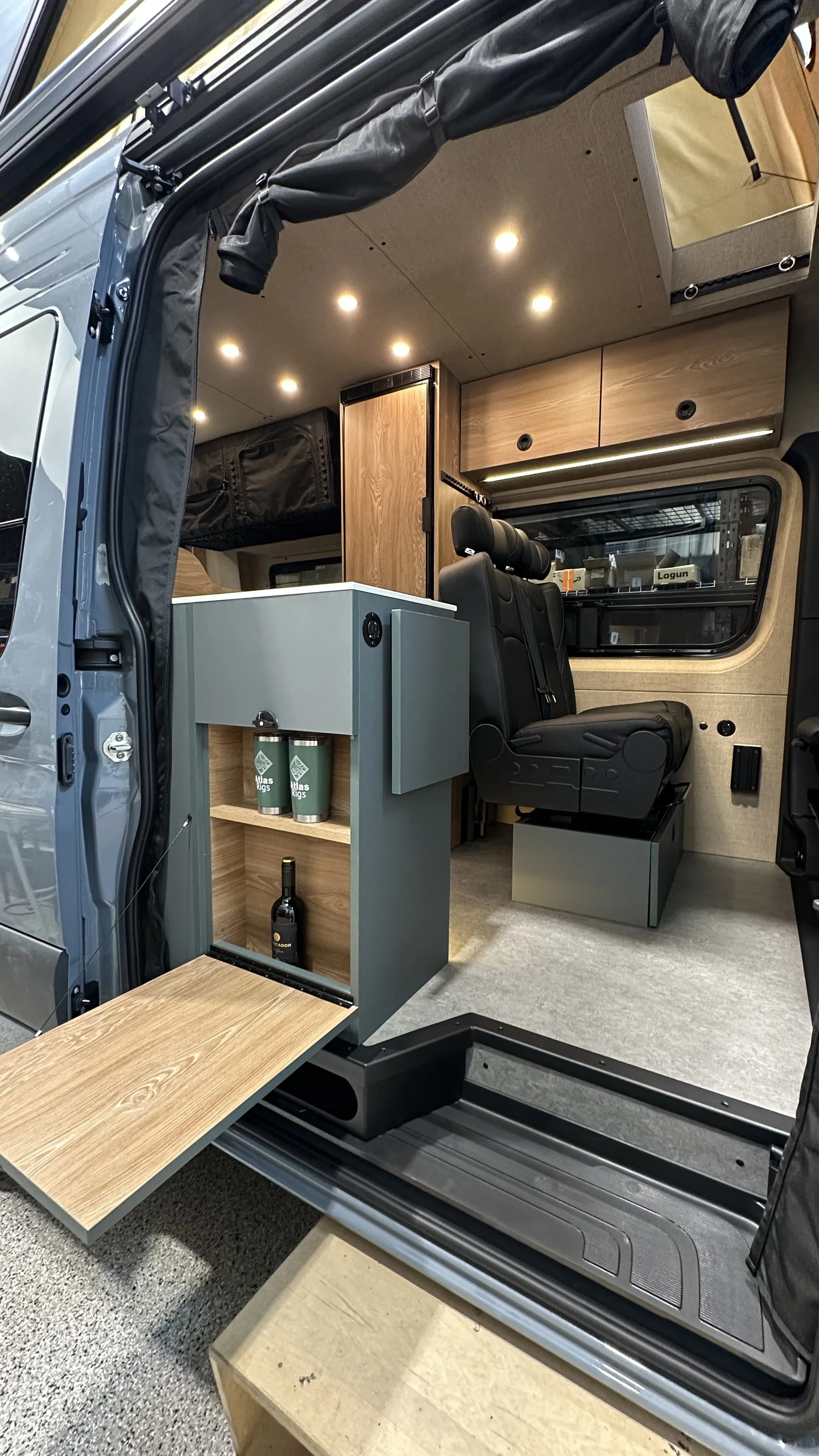 Interior of a camper van with wooden cabinets, two black seats, a large window, LED ceiling lights, a fold-out wooden table, and shelves with bottles and cups.