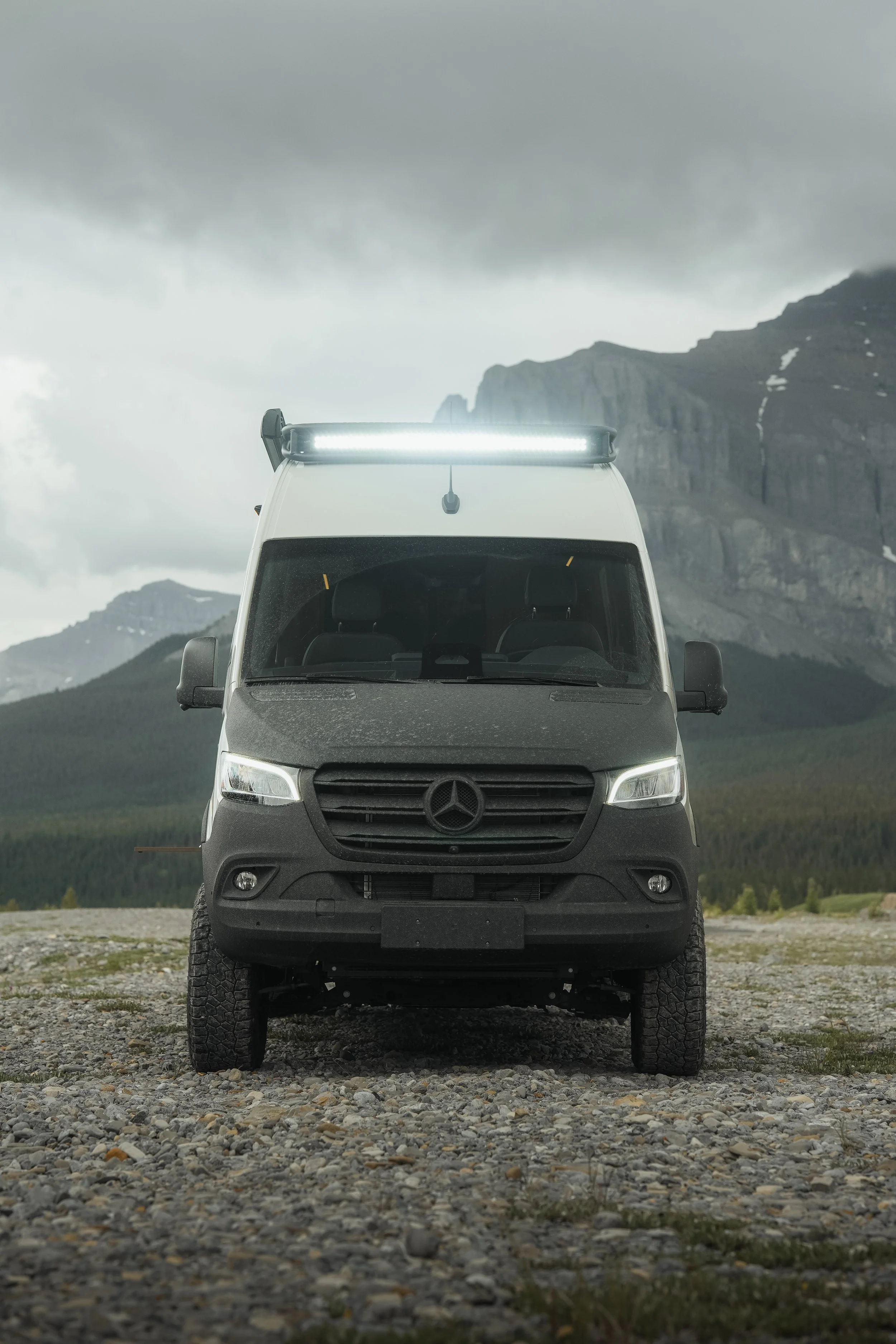 Front view of a white Mercedes-Benz van with a LED light bar on the roof, parked on rocky terrain with mountains and cloudy sky in the background.