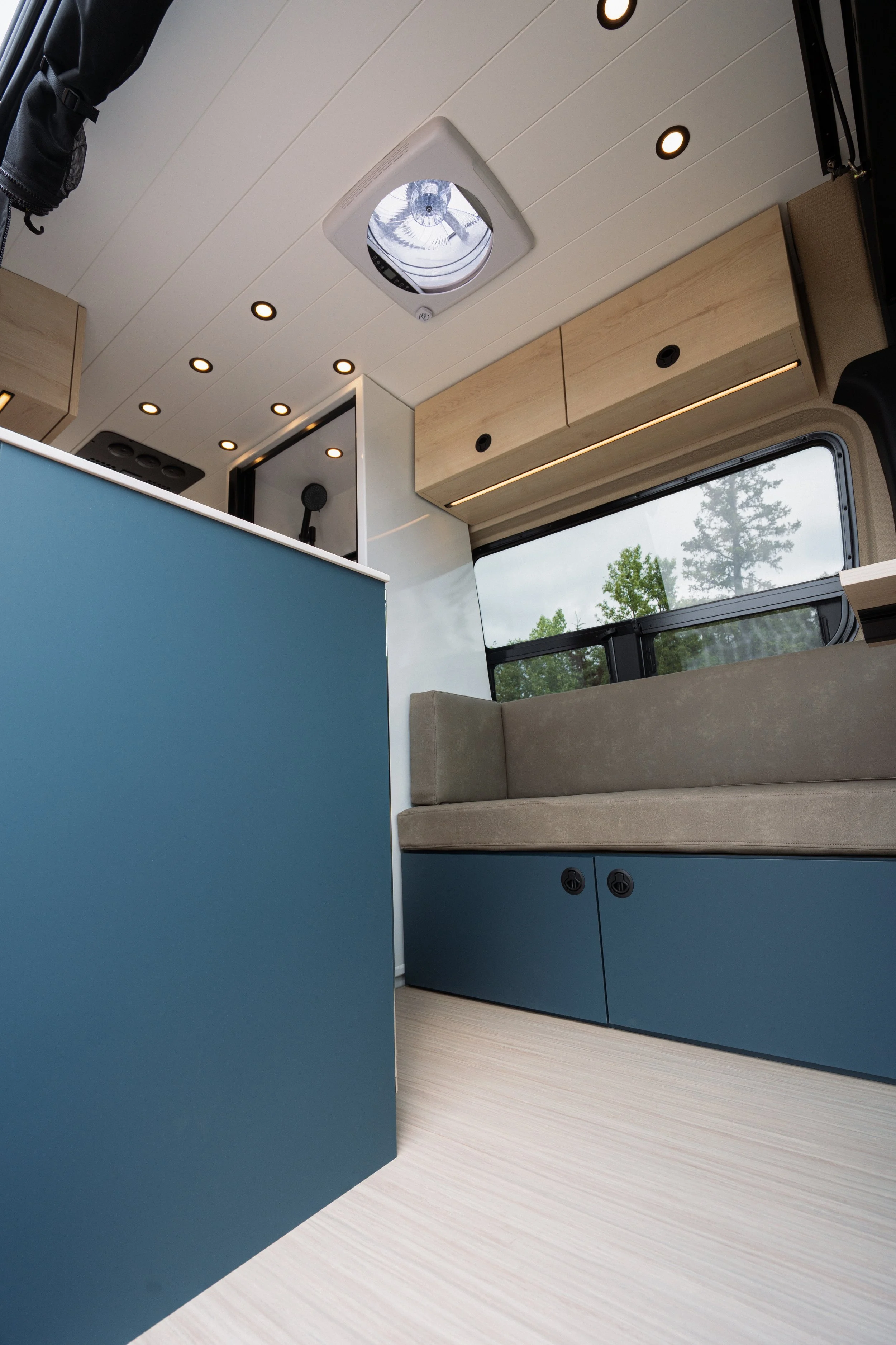 Interior of a modern camper or small living space with light wood cabinets, a window with a view of trees, and a ceiling with round recessed lights and a roof vent.