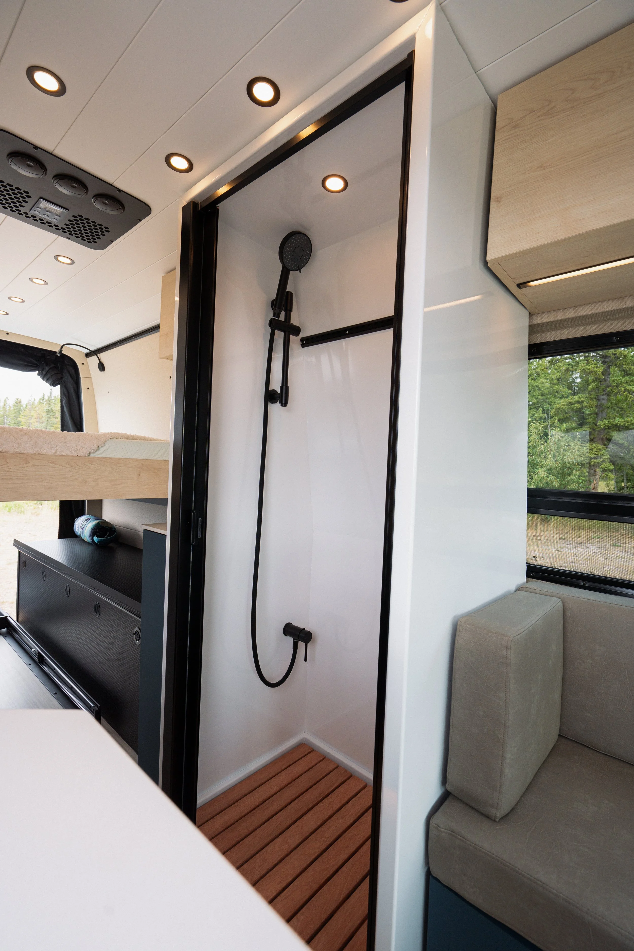 Interior of a camper or small living space with a shower area, a beige sofa, a window showing greenery outside, and ceiling lights.