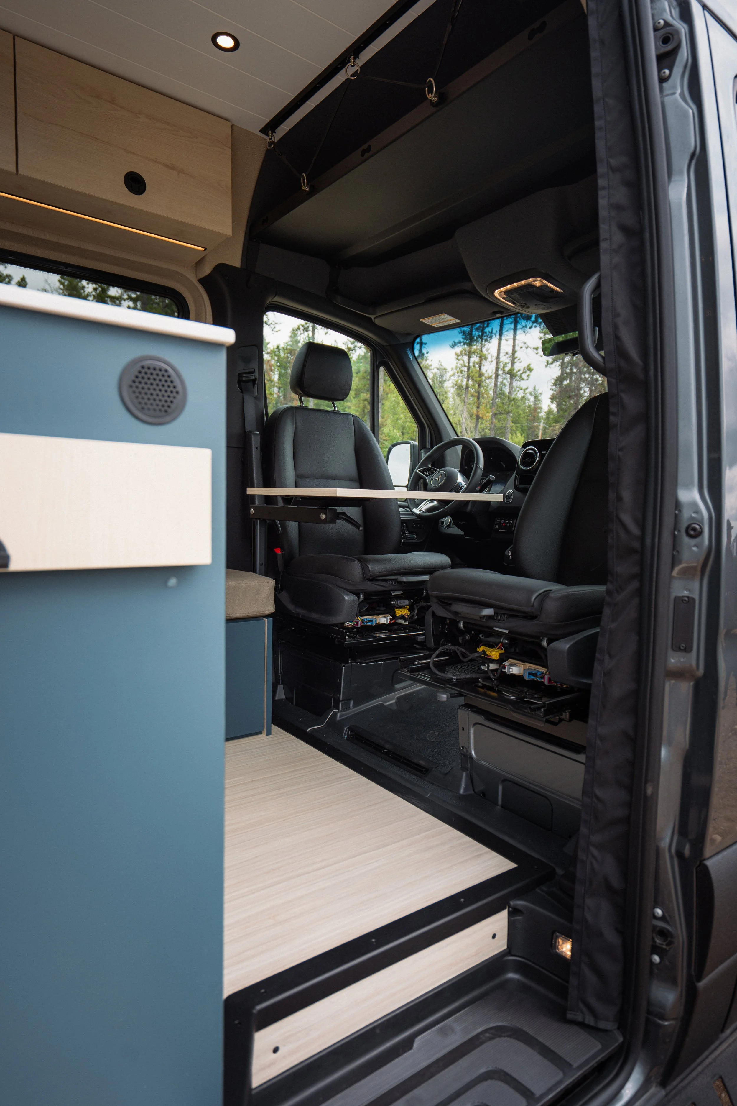 The interior of a van converted into a camper, with a bed platform, overhead storage, and a blue cabinet with a speaker, with a driver and passenger seat visible and a window showing trees outside.
