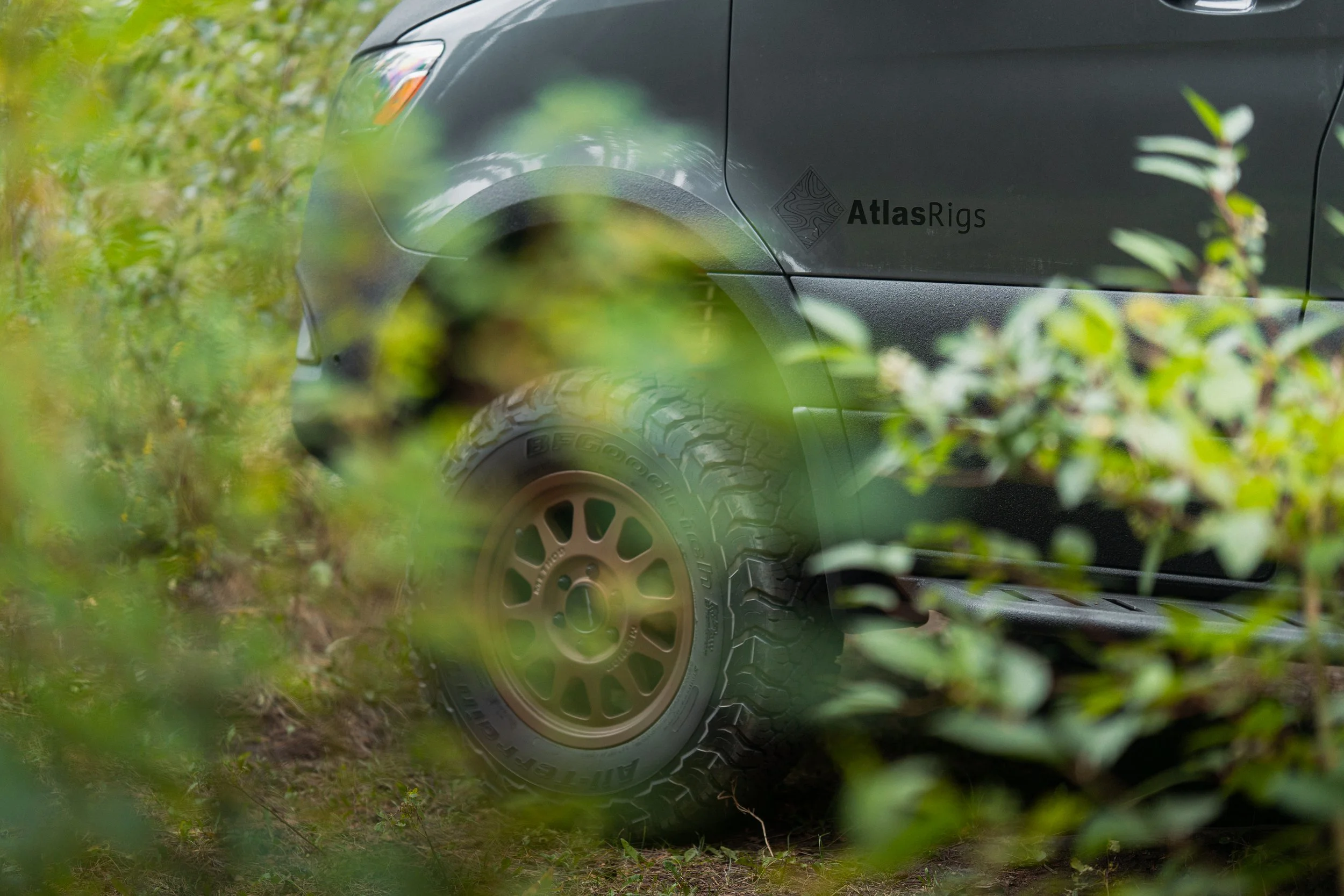 A gray vehicle partially visible behind green foliage, with a badge that says 'Atlas Rigs' on the side, parked on ground with grass and dirt.
