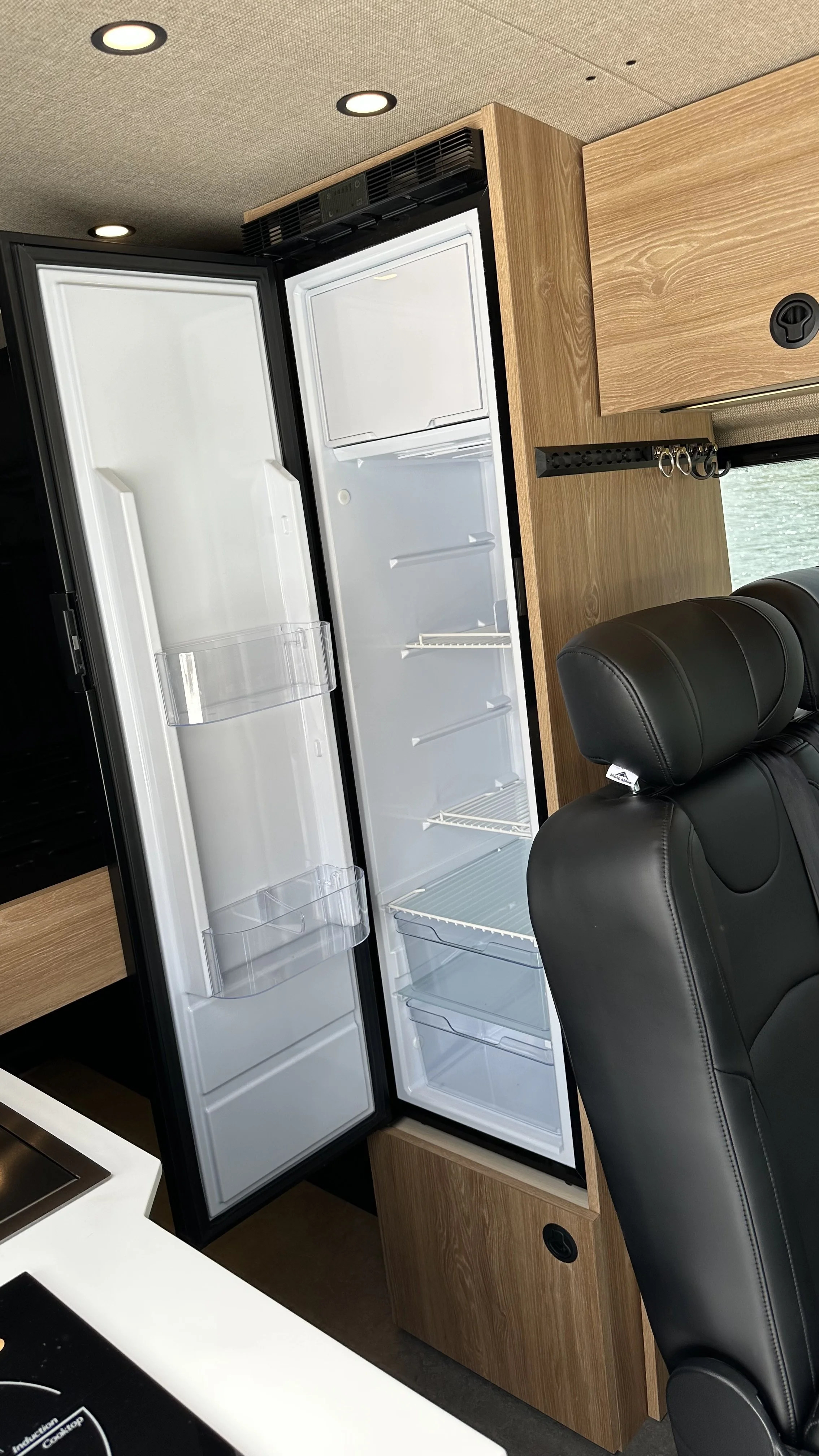 Open black refrigerator with empty shelves and door compartments next to a black leather chair and wooden cabinet in a kitchen area.