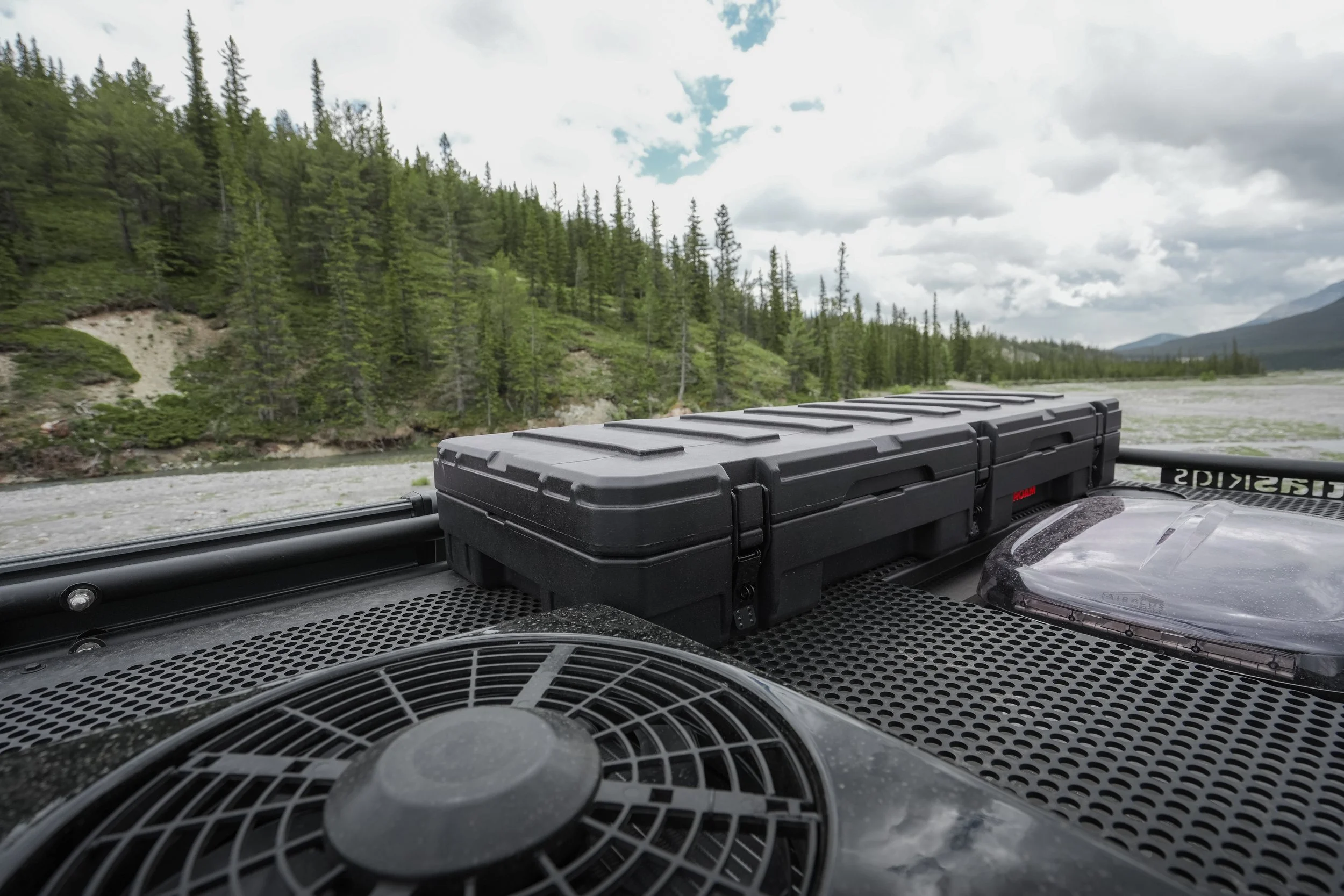 View from inside a vehicle showing a black storage box and other equipment on the dashboard with a scenic view of a river, green pine trees, and cloudy sky in the background.