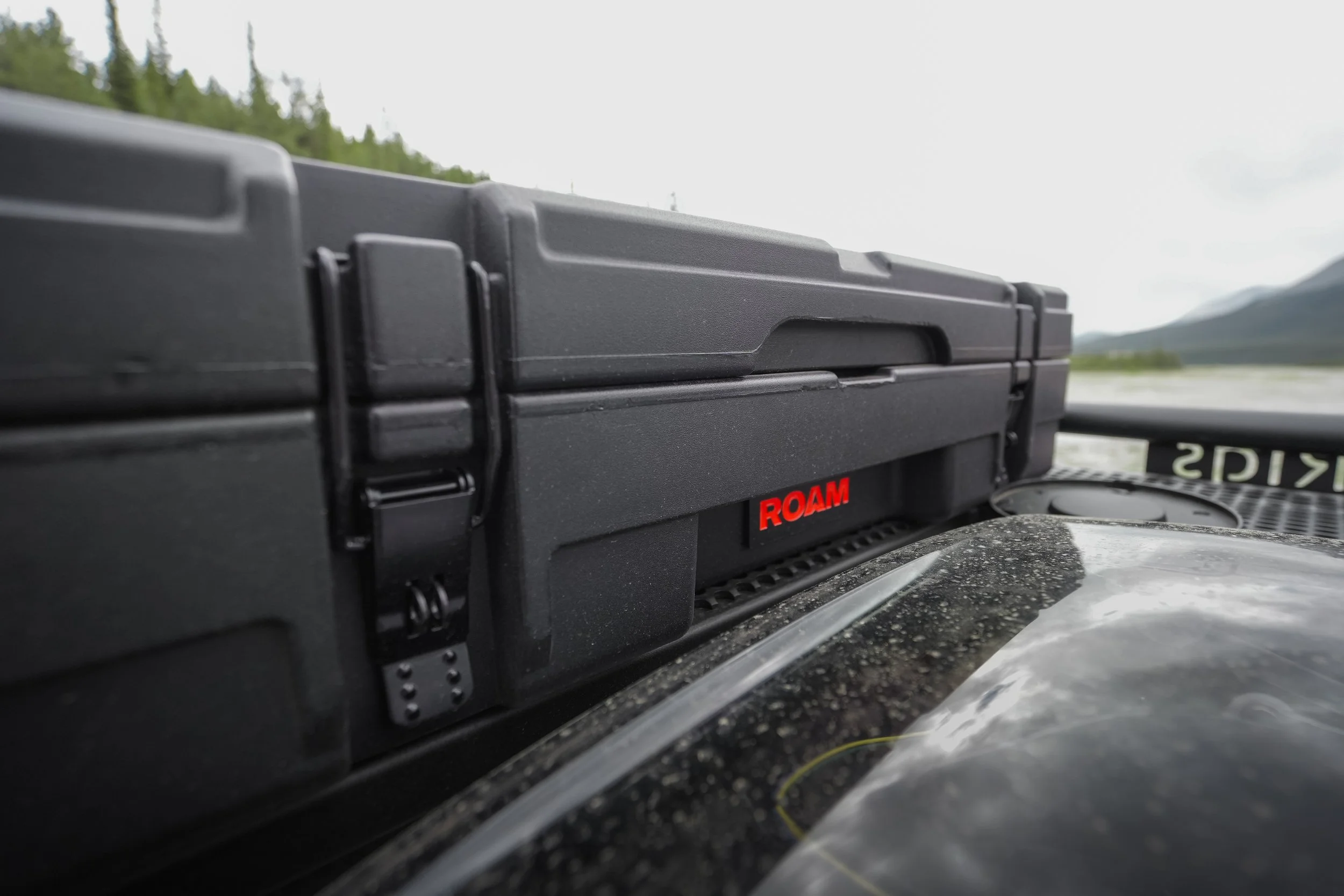 Close-up of a black ROAM storage case placed on a vehicle dashboard with mountainous landscape and cloudy sky in the background.