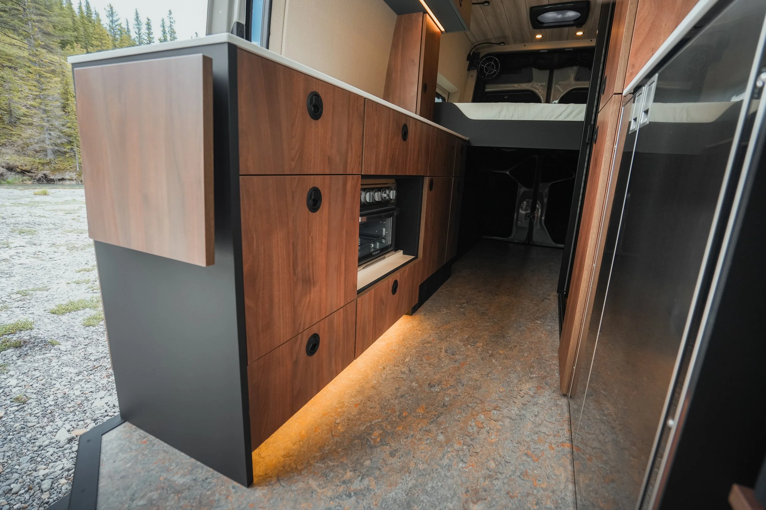 Interior of a camper van showing wooden cabinets, a countertop, and a stove with a window overlooking a river and trees outside.