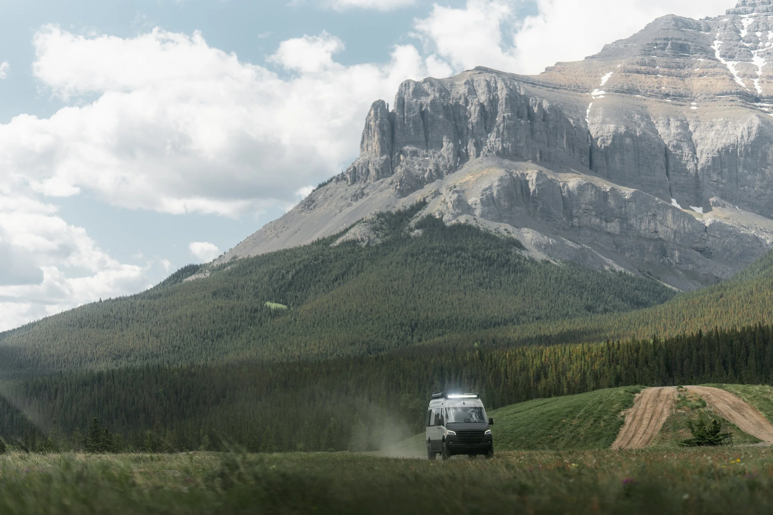 A mountain with rocky cliffs and patches of snow, lush green forest at its base, cloudy sky, and a black van driving on a dirt road in front of a cultivated field.