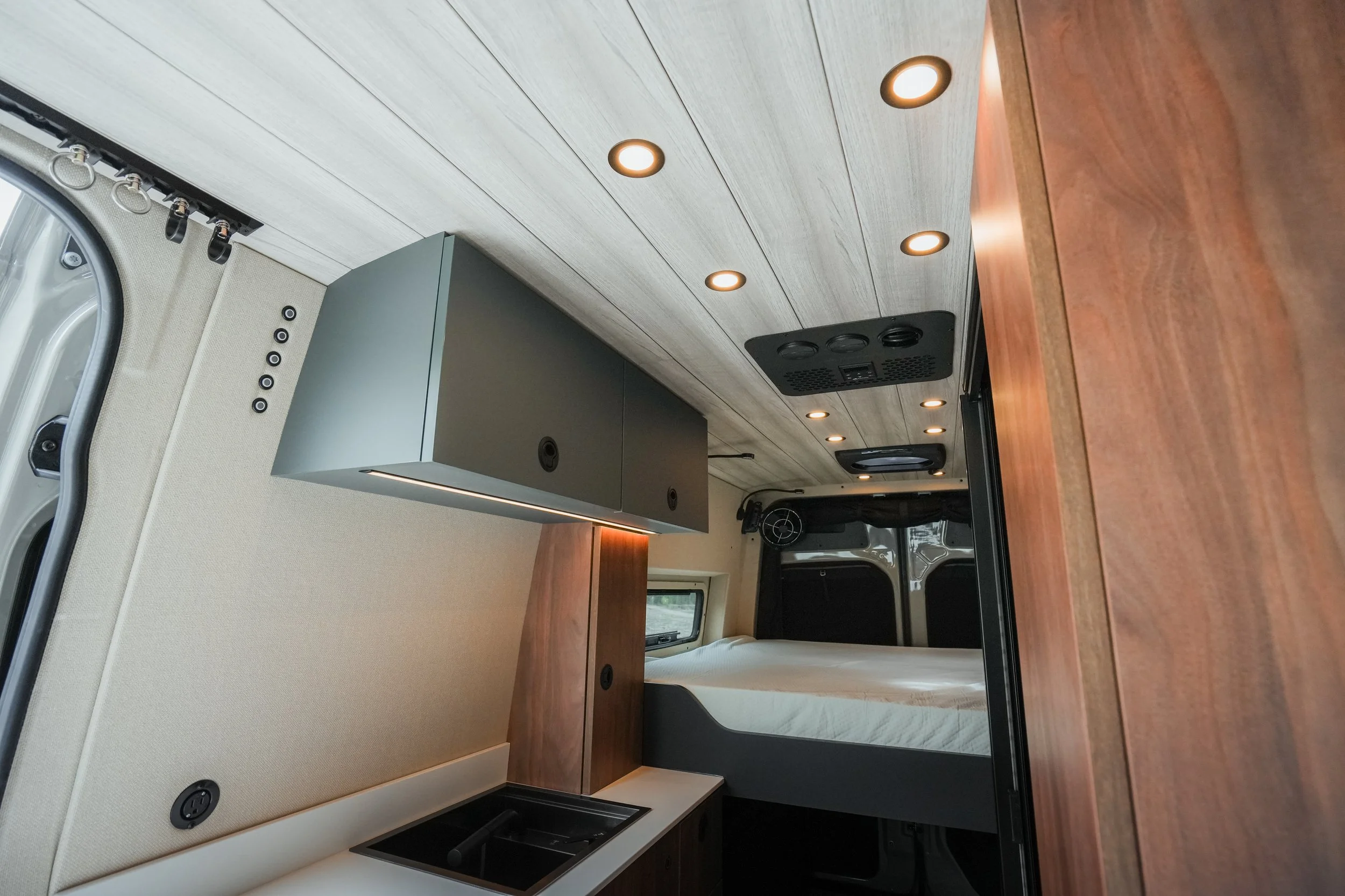 Interior of a camper van with wooden countertops, a small black sink, a gray cabinet, beige walls, and recessed ceiling lights.