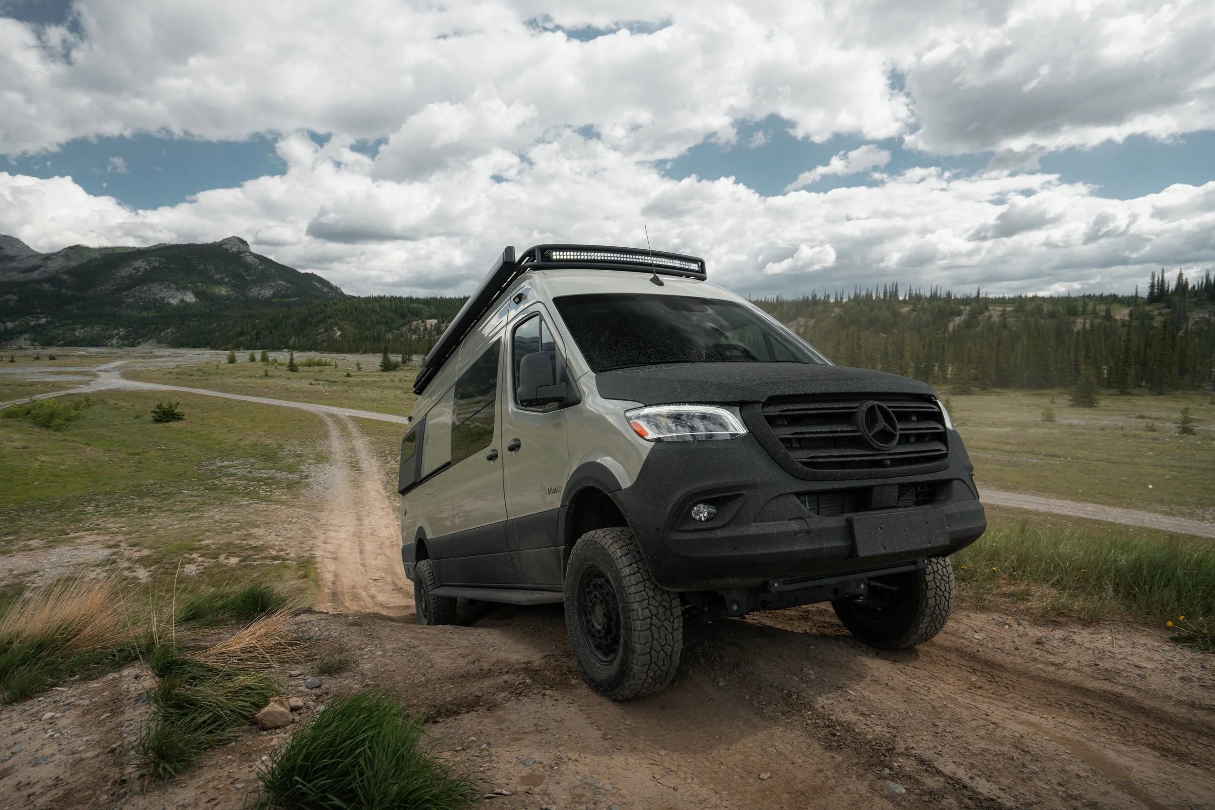 A white off-road van driving on a dirt trail through a grassy field with mountains and a partly cloudy sky in the background.