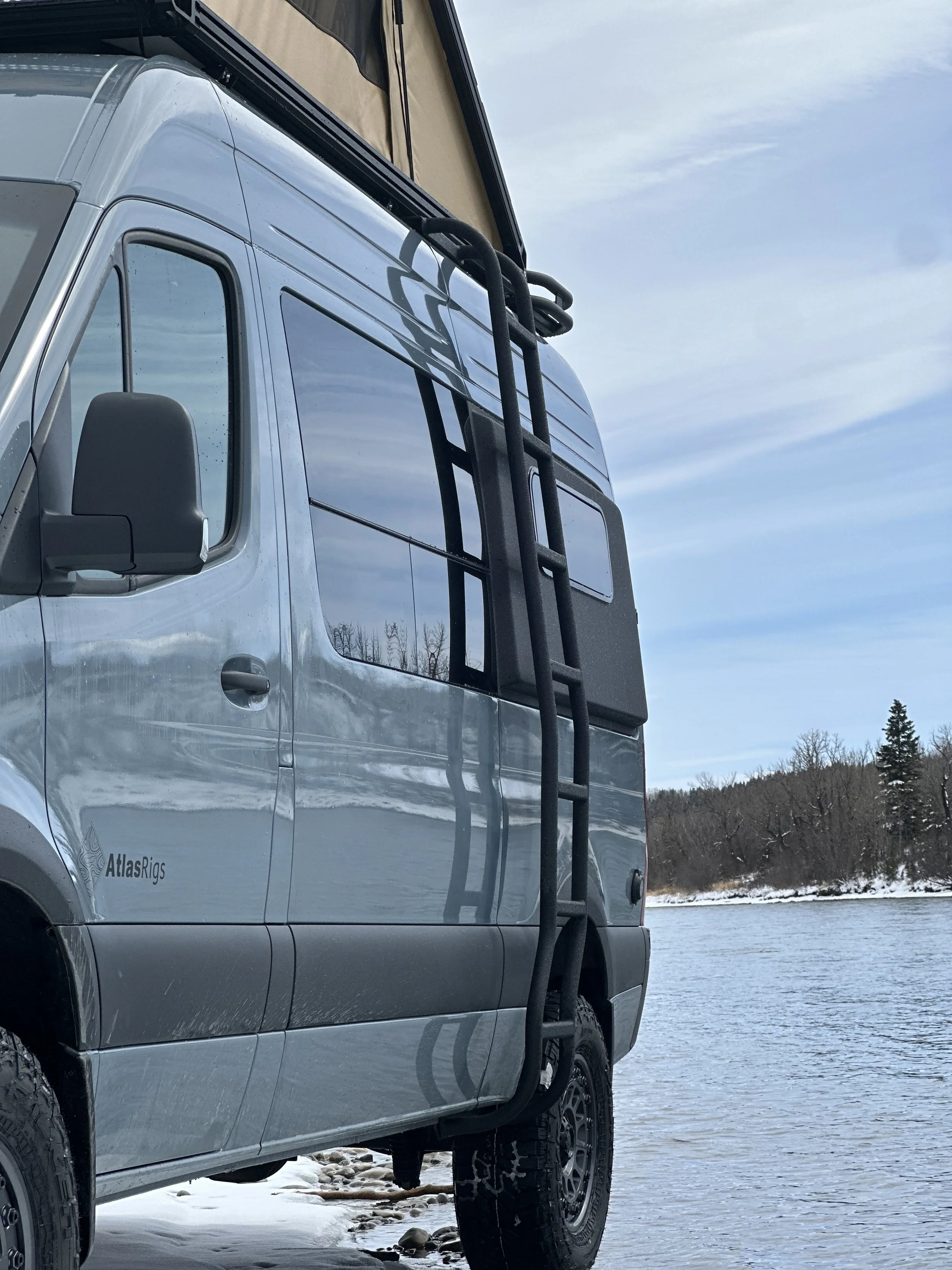 Close-up of a silver pickup truck with a ladder attached to its side, parked by a frozen lake or river with snow on the ground and bare trees in the background.