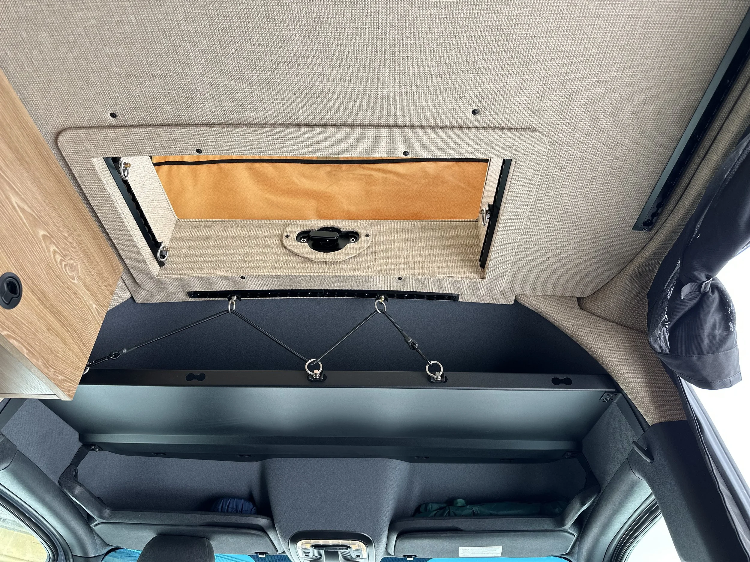 The interior ceiling of a vehicle with a rectangular hatch, black handles, a circular access port, and metallic fixtures.