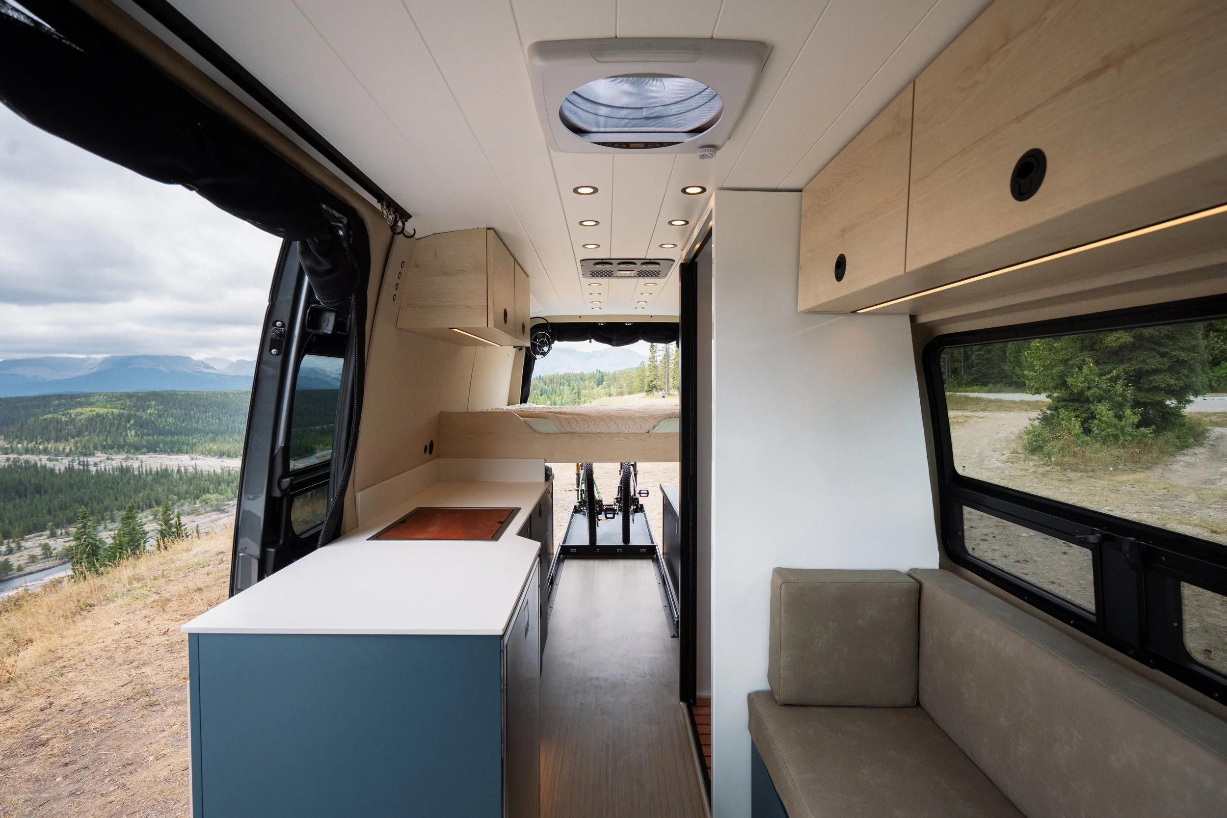 Interior view of a camper van with a kitchen area, seating, and large windows showing a scenic outdoor landscape of trees, mountains, and a cloudy sky.