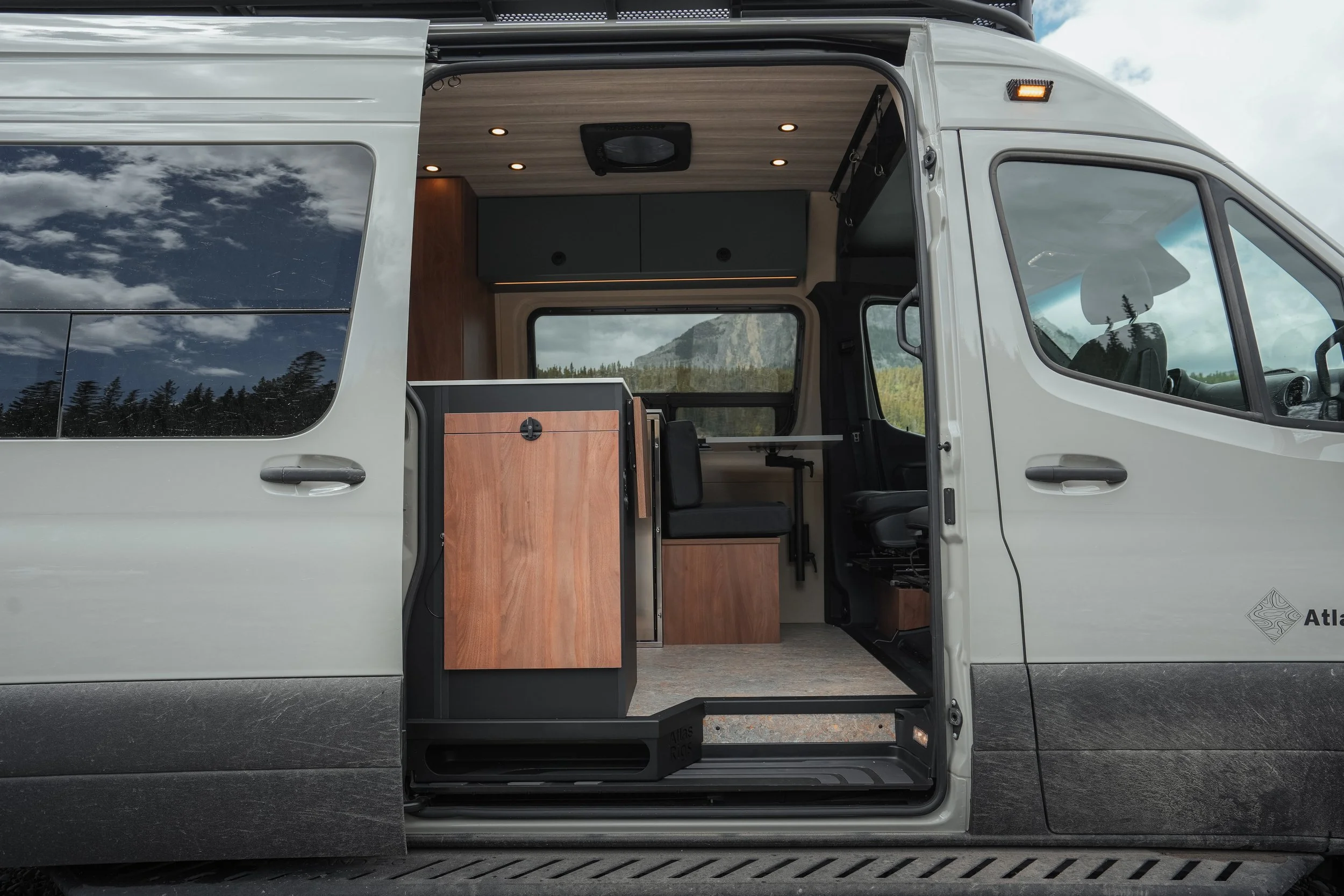 Interior of a white camper van with wooden cabinetry, black overhead storage, and a window showing a scenic mountain landscape.