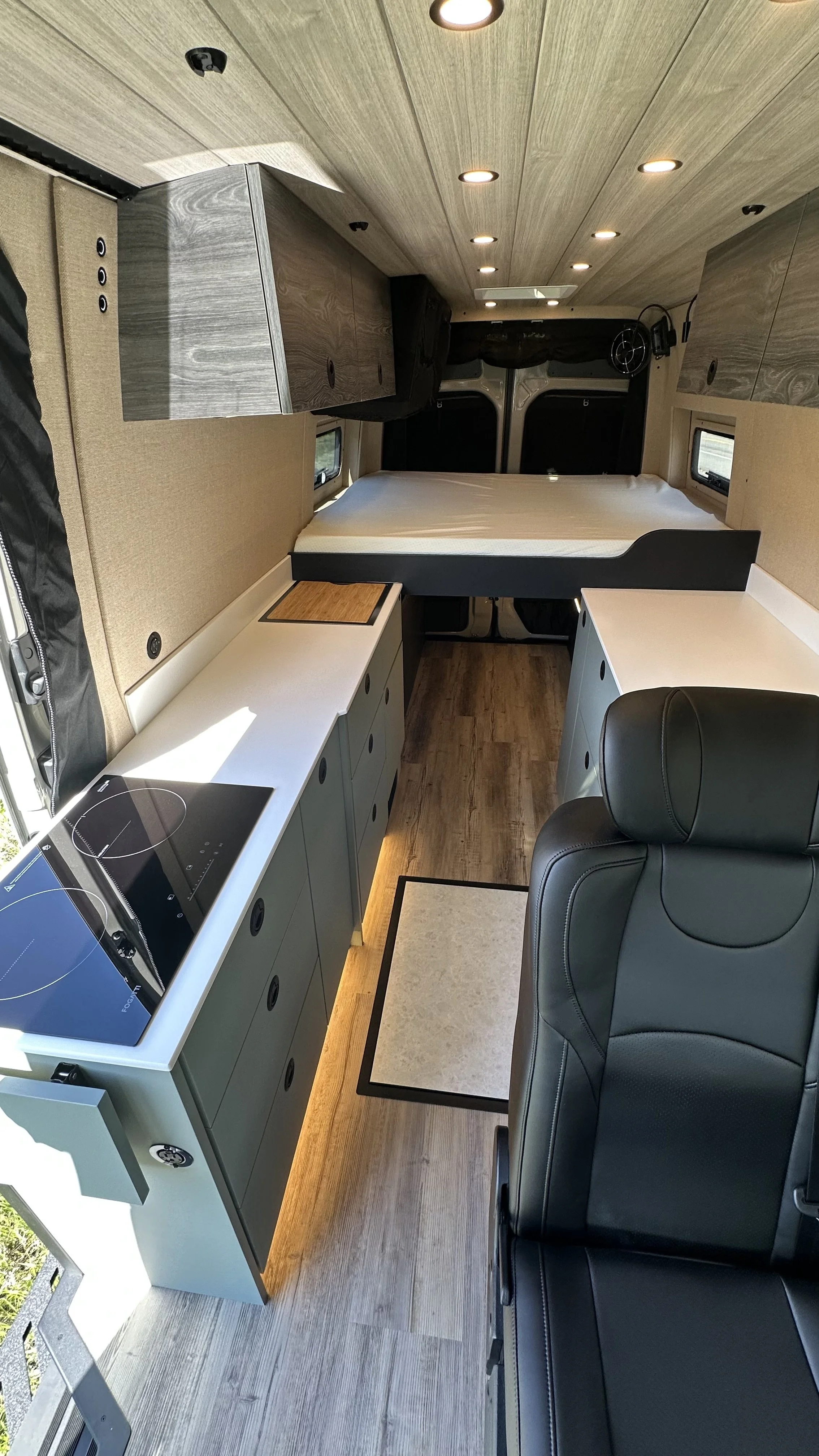 Interior of a modern camper van or RV featuring a bed, kitchen area with cooktop, storage cabinets, a black leather seating area, and small windows, all within a compact space with wood and neutral-colored finishes.