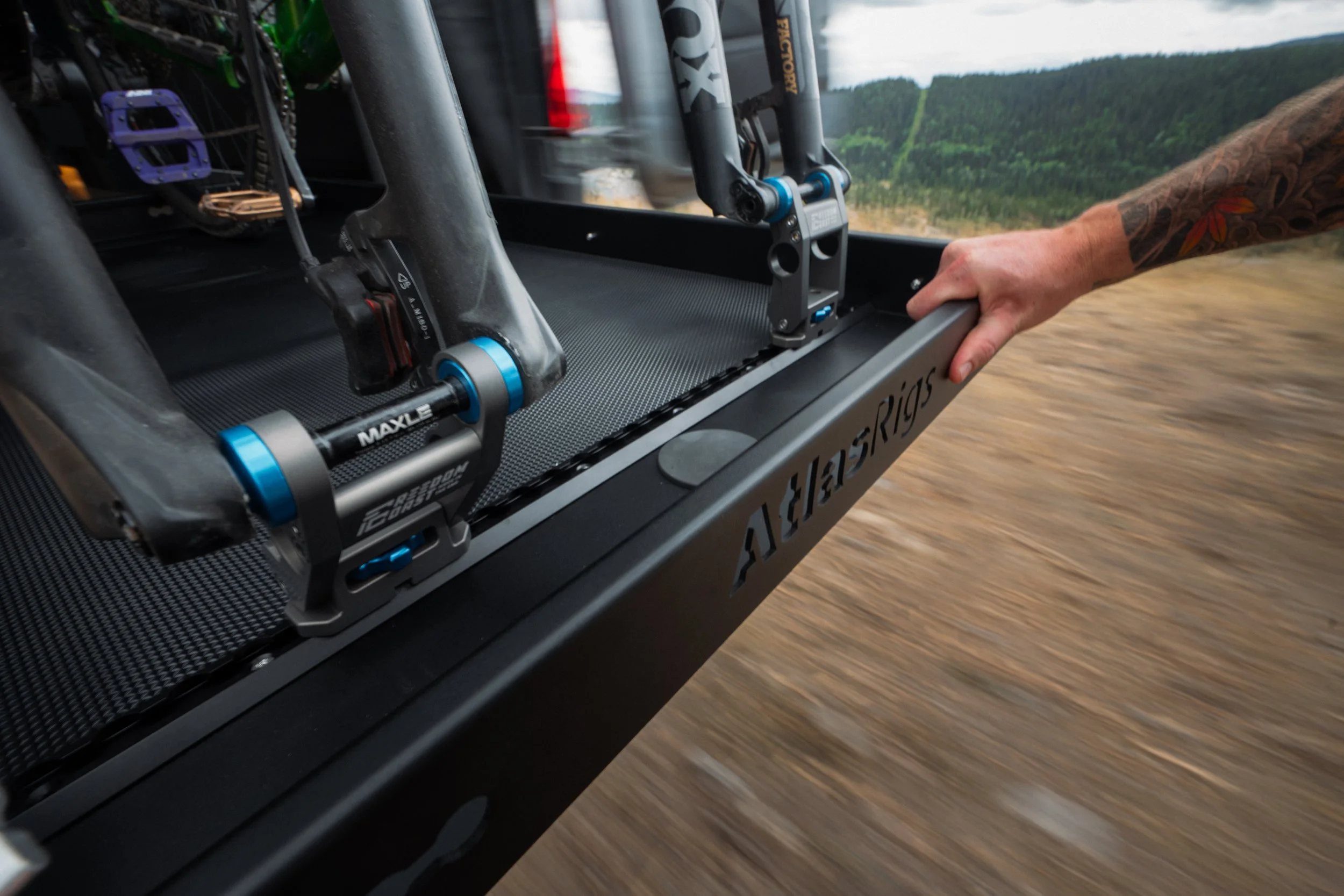 Close-up view of a mountain bike suspension fork and components mounted on a small vehicle or cart, traveling on a dirt trail through a forested landscape with green trees and hills in the background.