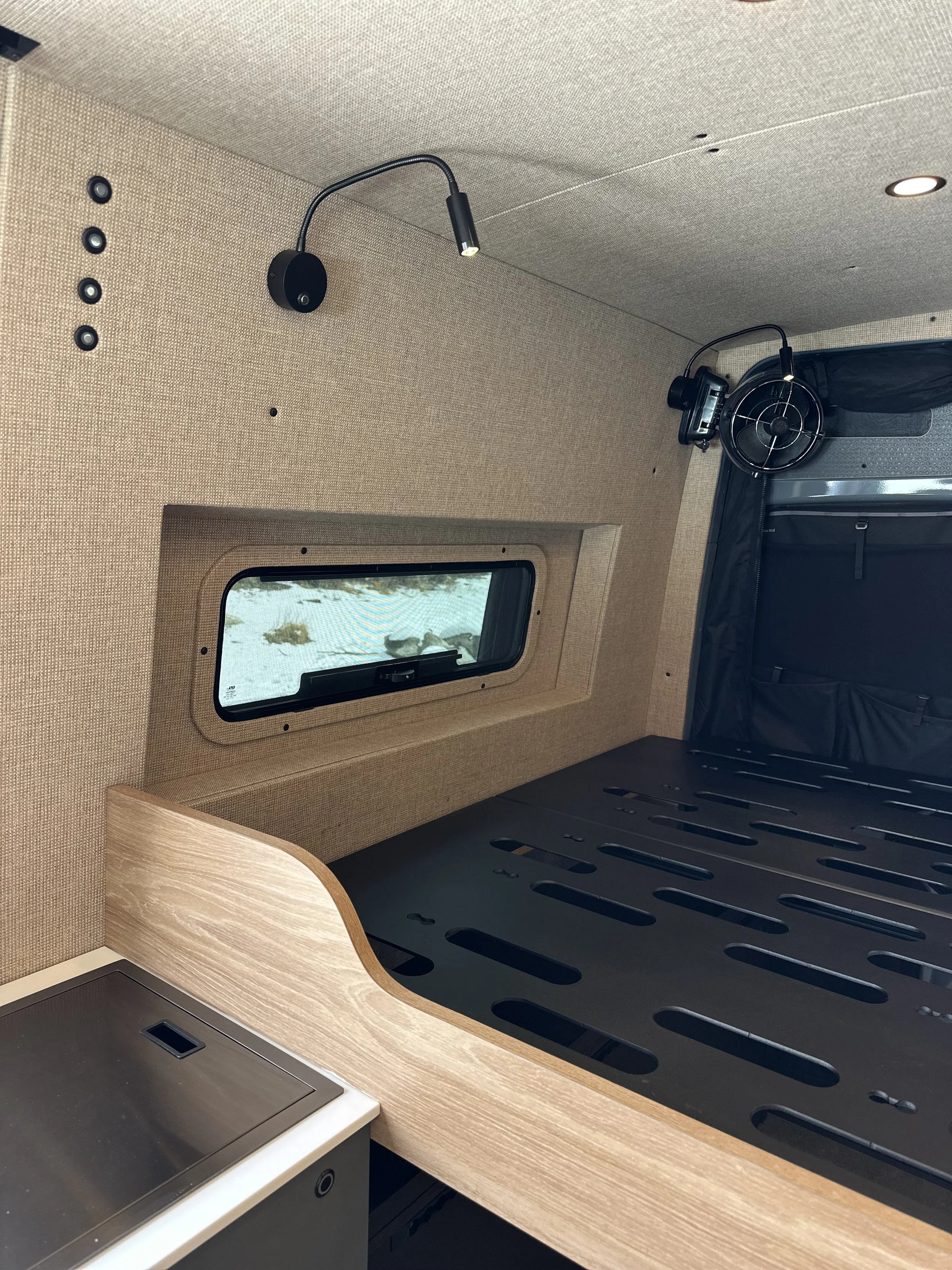 Interior of a compact kitchen or workspace with a small window, beige textured walls, black countertop with a cutout, and a black rack or stove underneath. There are two black wall-mounted LED lights and a black camera or sensor on the wall.