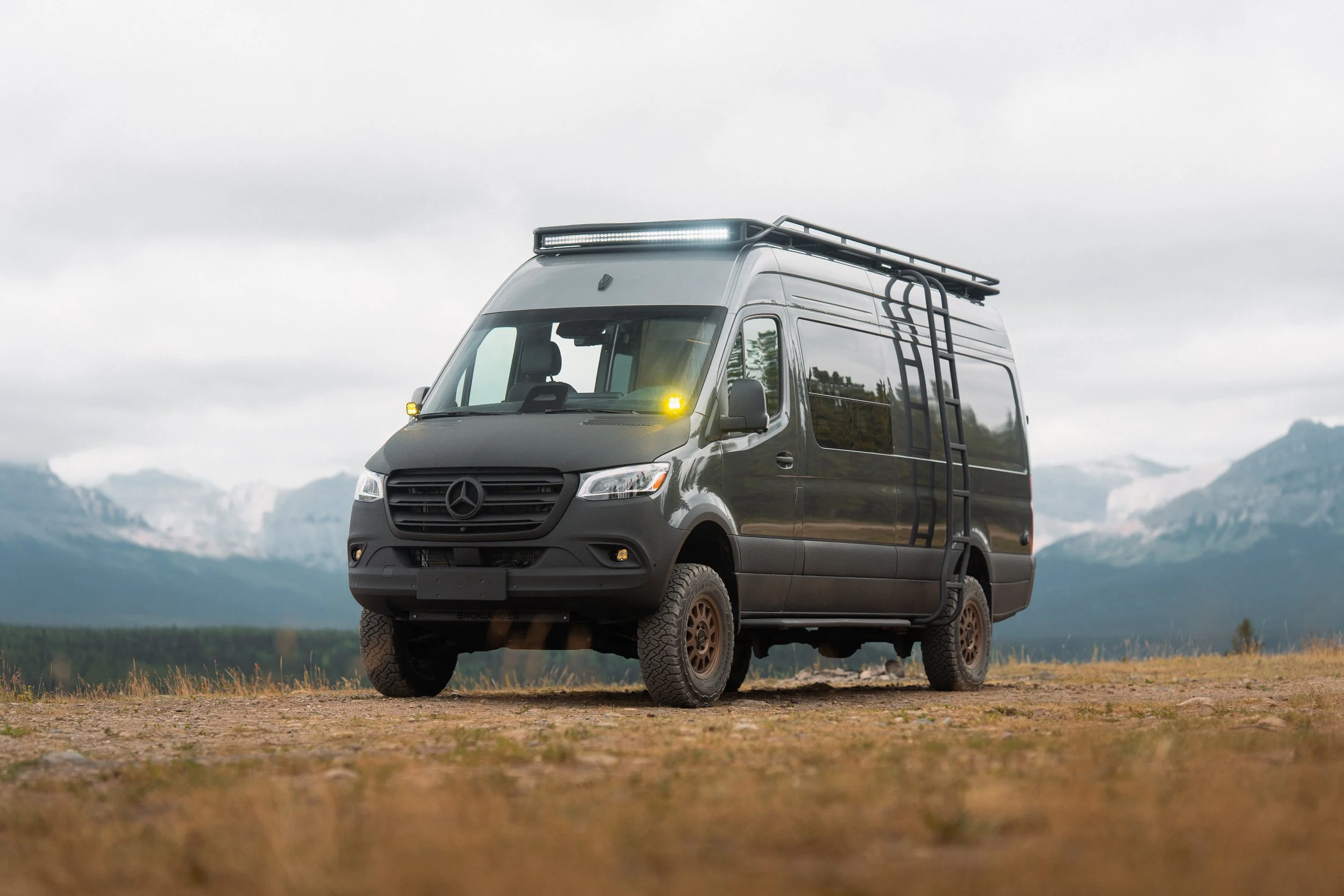A black camper van with off-road tires parked in a grassy field with mountains and cloudy sky in the background.