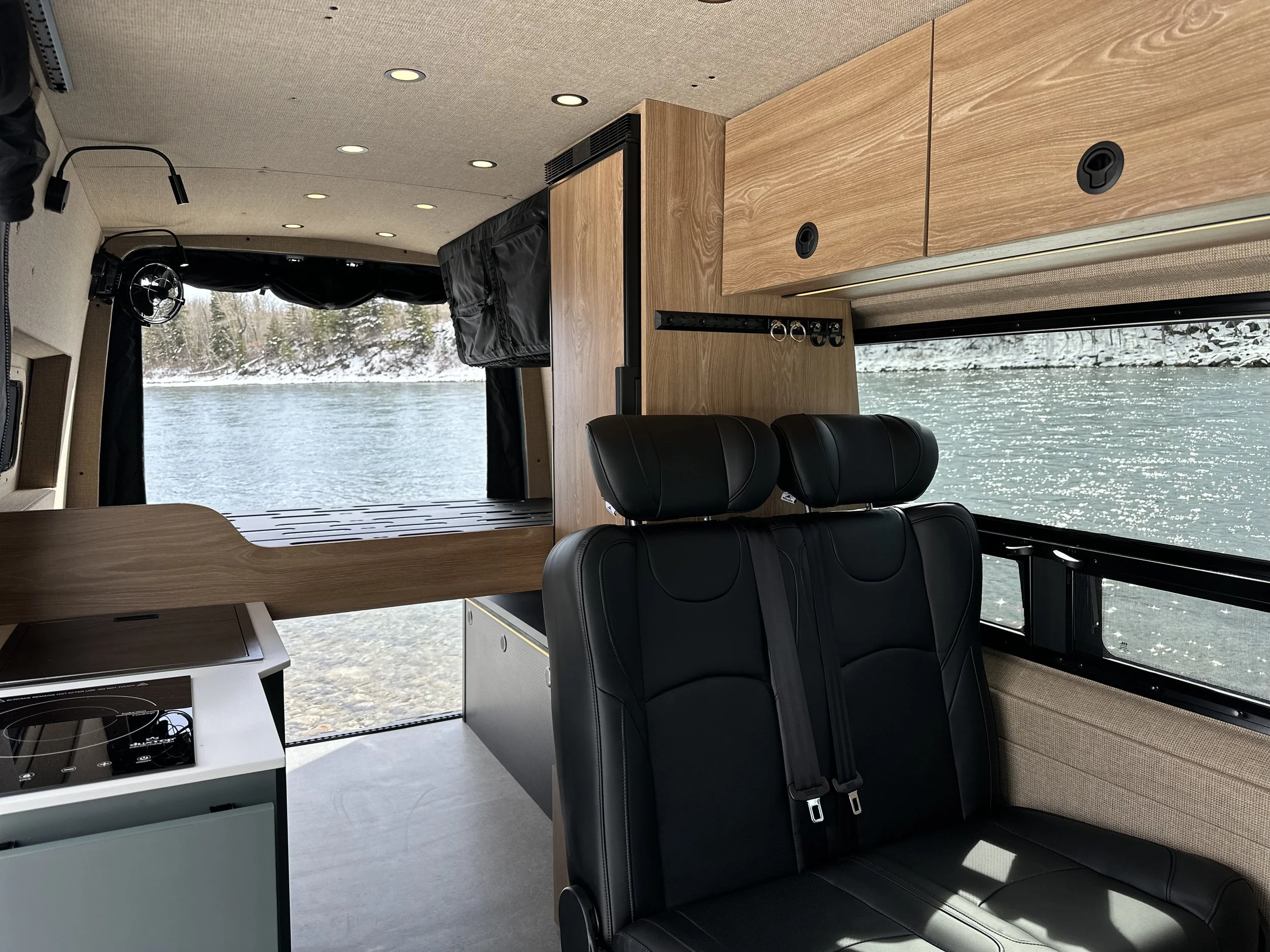 Interior of a camper van with black leather seats, wooden cabinets, a small stove, and a large window showing a snowy landscape with trees and water.