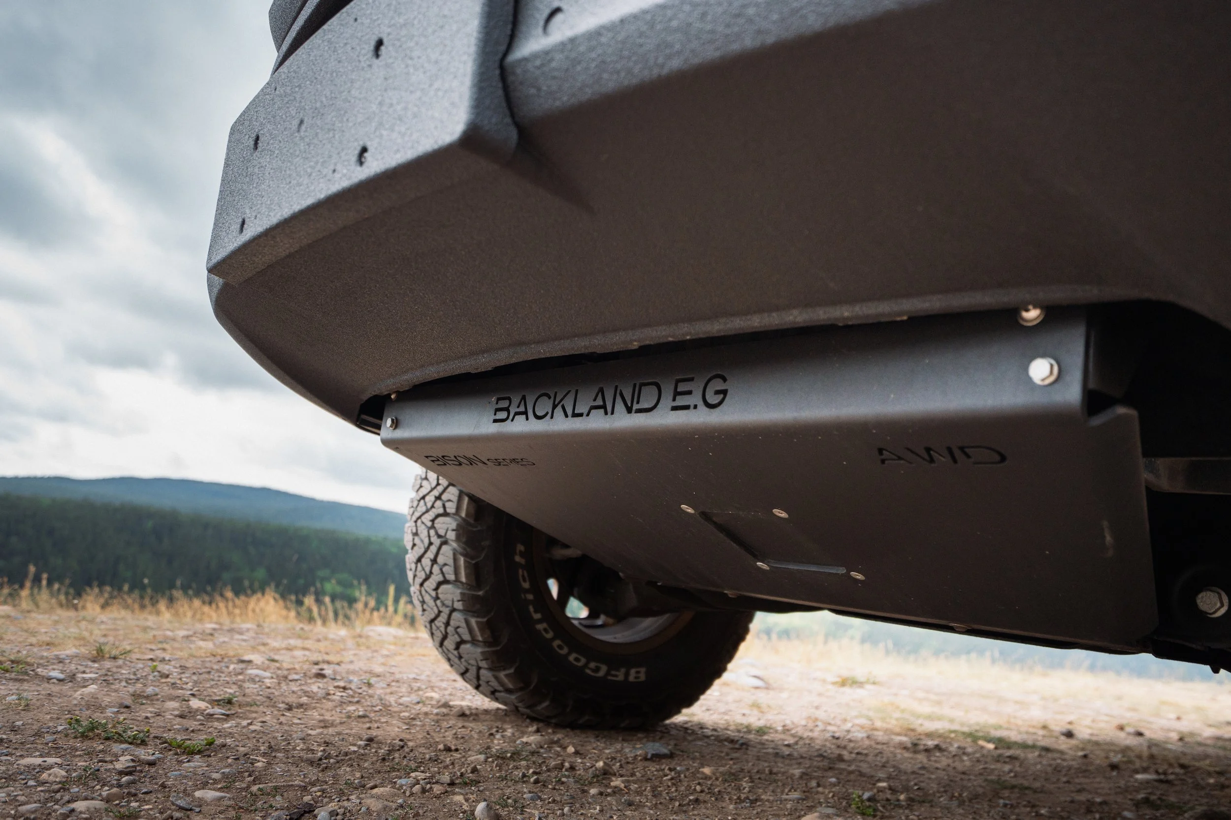 Close-up of the underside of a vehicle, showing the tire and the metal skid plate with the text "BACKLAND E.G" on it, in an outdoor setting with mountains and cloudy sky in the background.