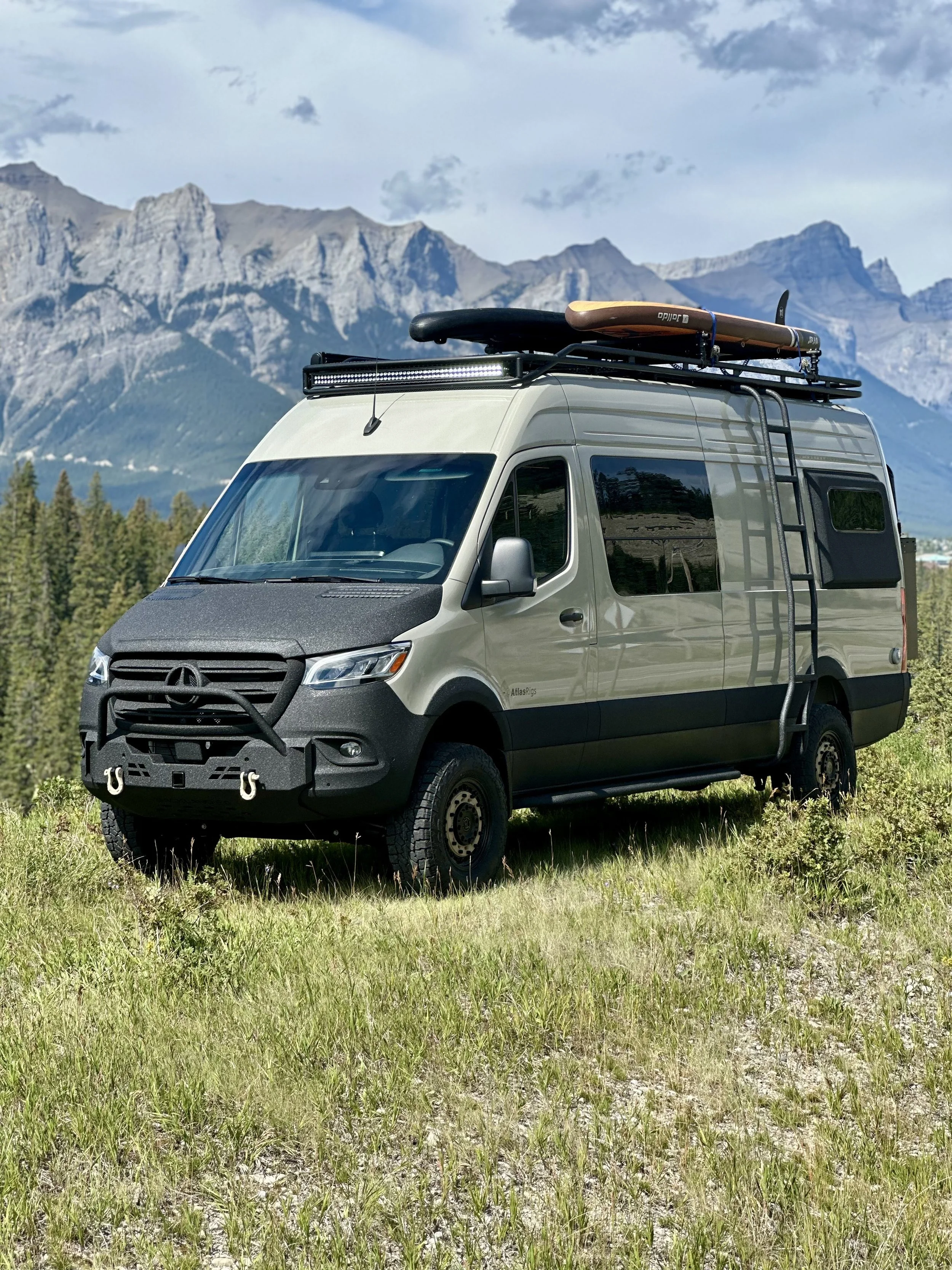 A beige camper van parked on green grass in a mountainous area with rocky peaks and a cloudy sky in the background. The van has a ladder on the side, a rooftop cargo rack with a surfboard, and some equipment mounted on top.